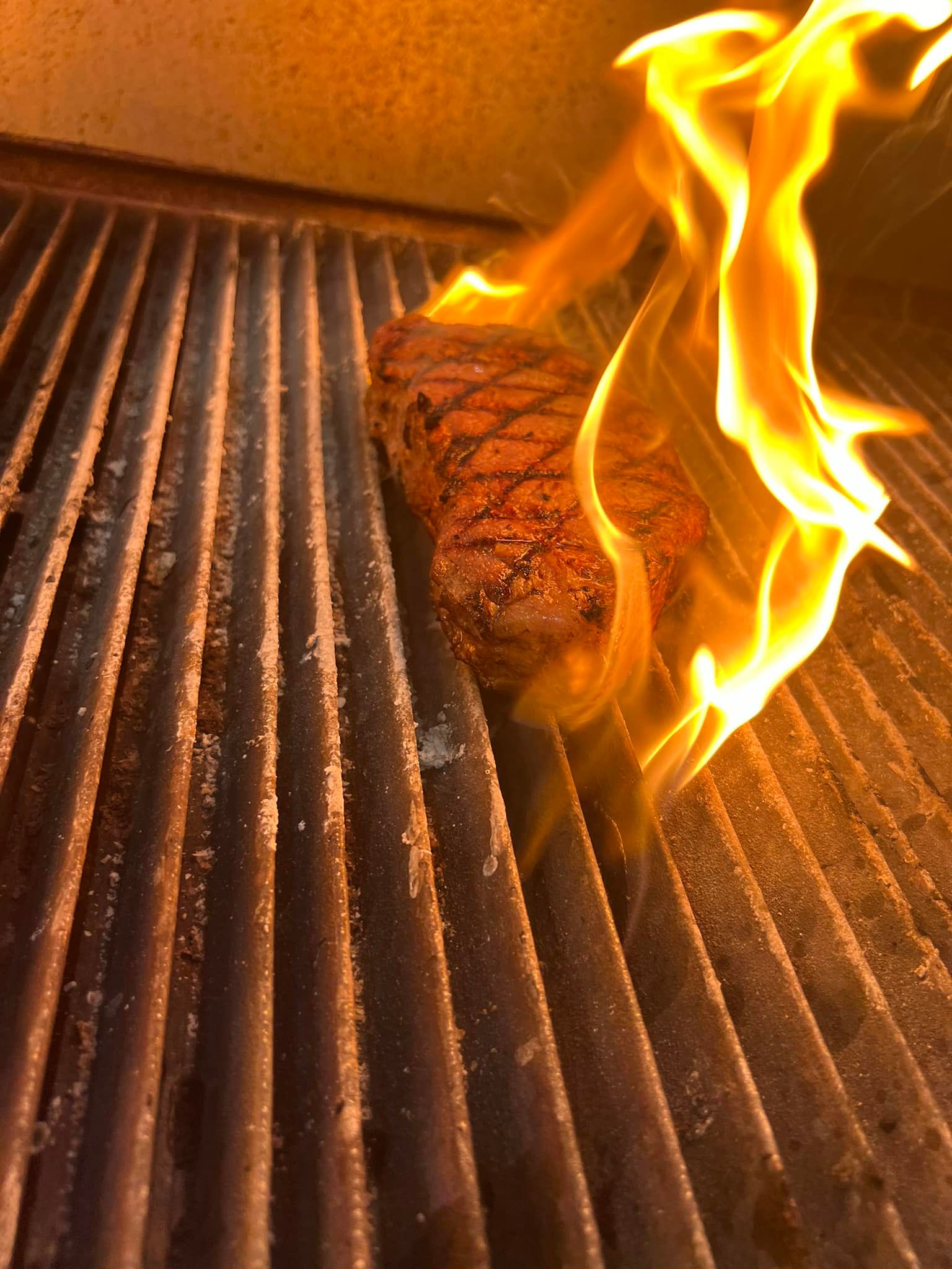 A seasoned steak cooking on a hot grill with prominent flames licking the side of the meat.