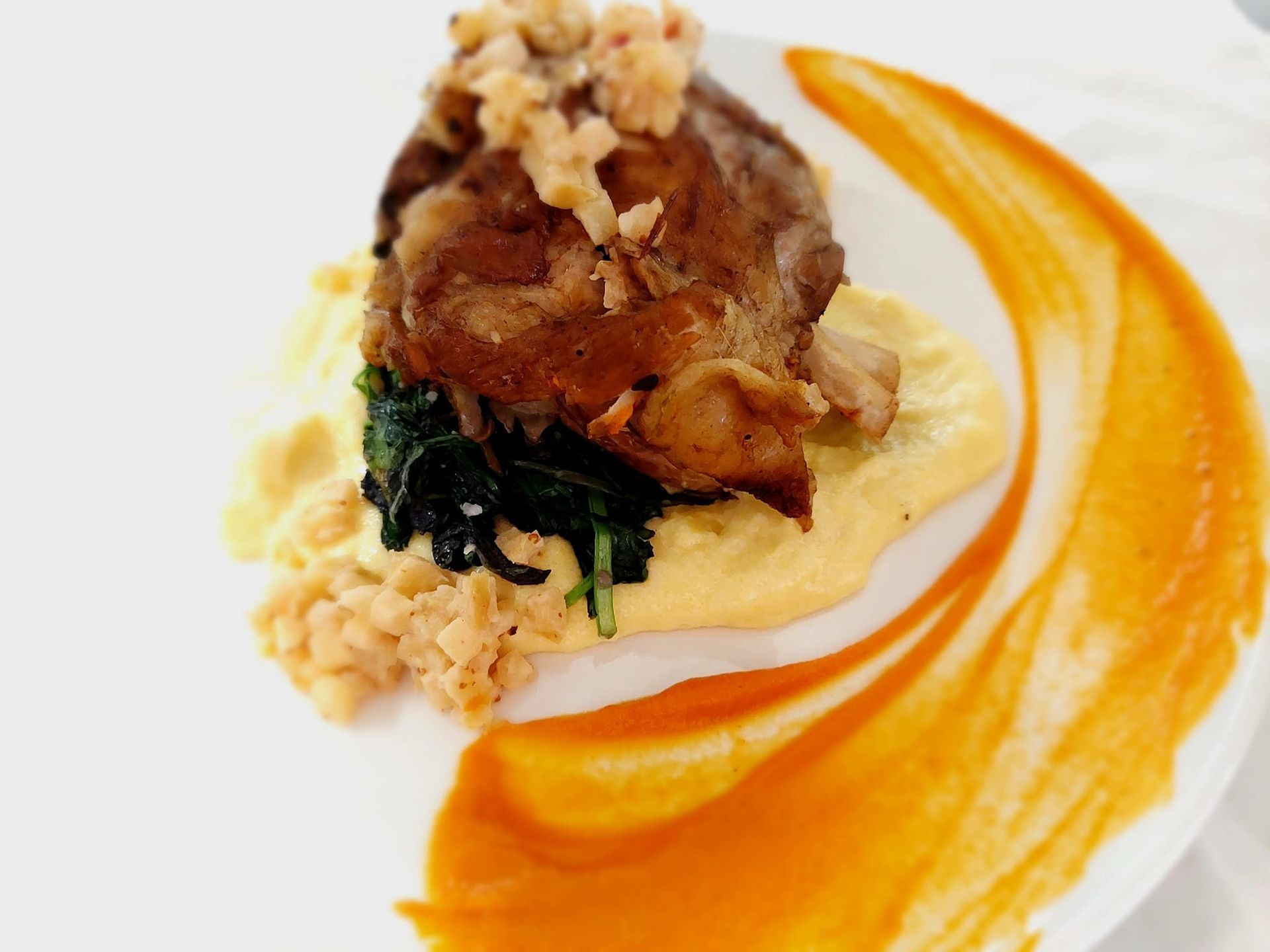 A cooked meat portion sits on purée and dark greens, garnished with a crumble, with a curved orange sauce swirl alongside.