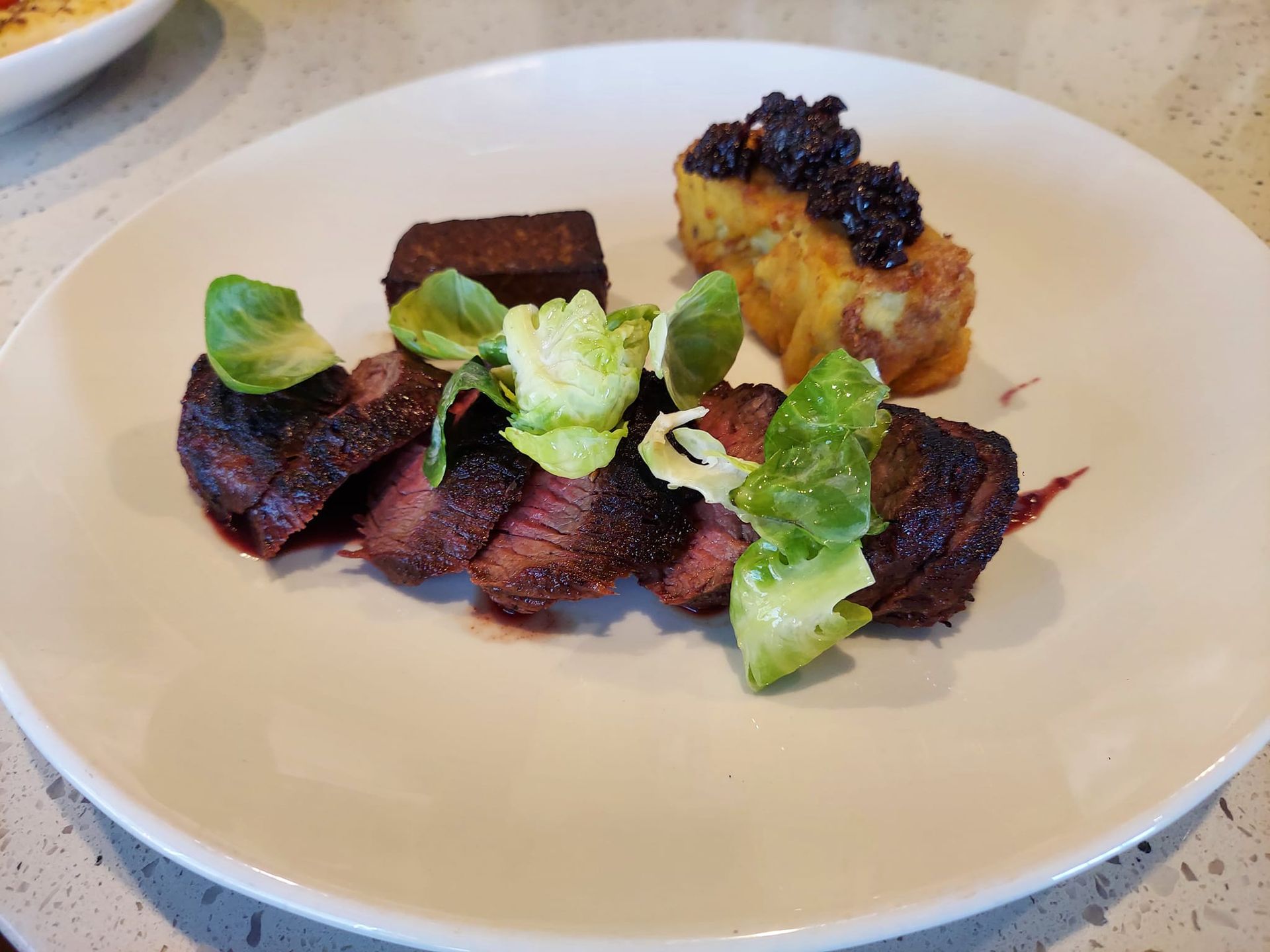 A plated dish featuring sliced, seared meat topped with small, bright green leafy greens, served with a potato square.