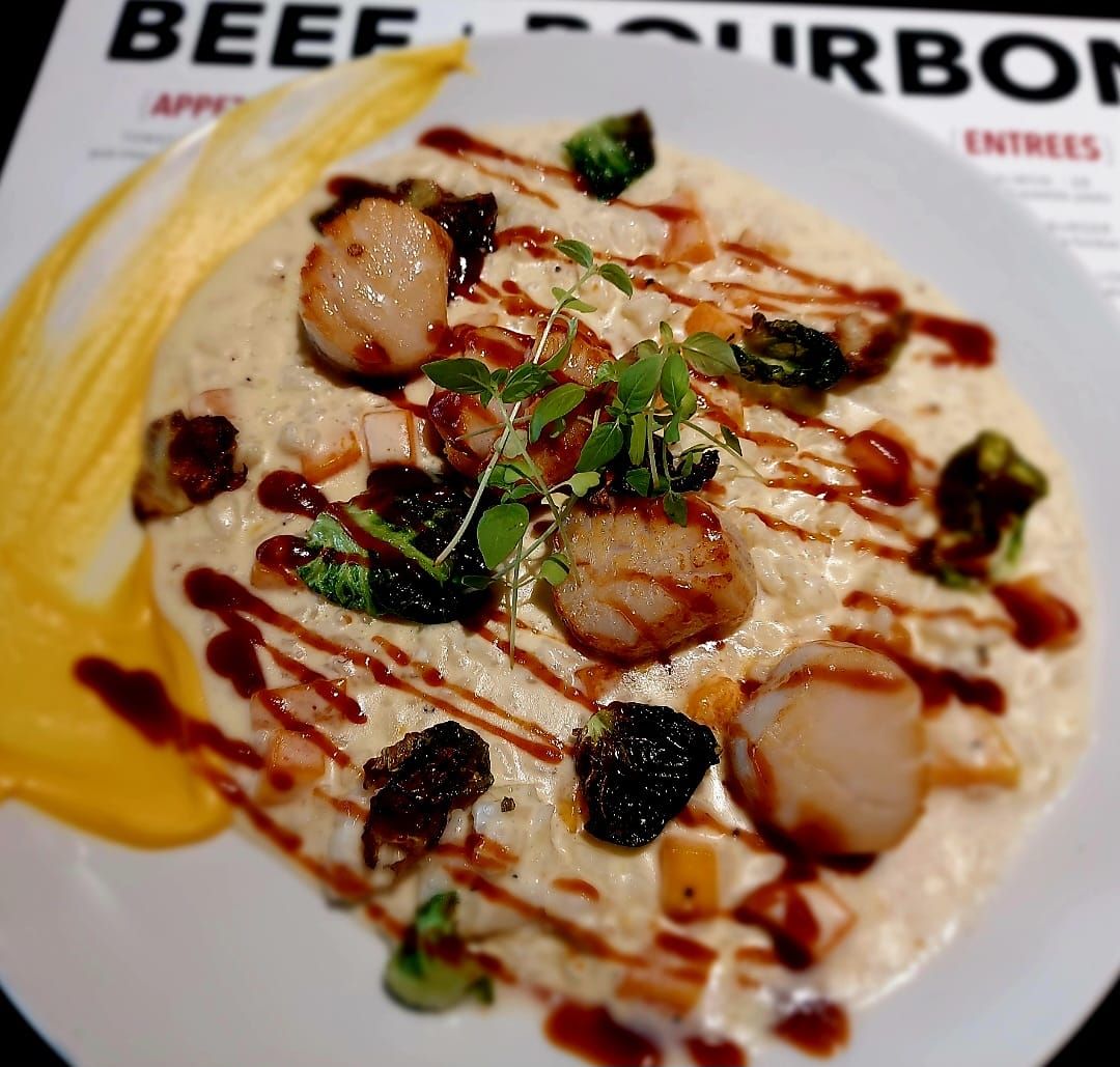 A plate of creamy risotto topped with seared scallops, roasted Brussels sprouts, and a savory drizzle, near a menu.
