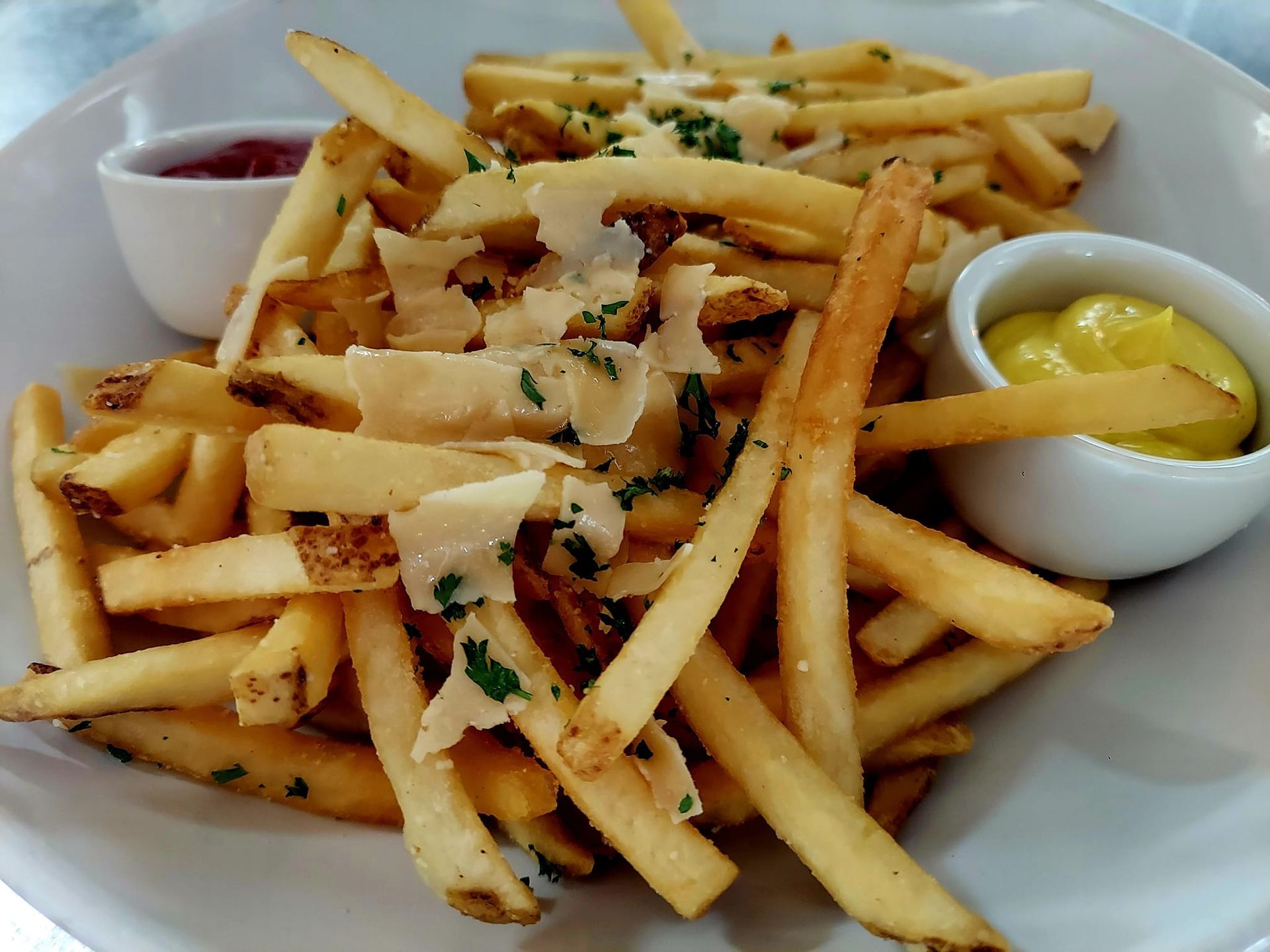 A bowl of golden-brown french fries topped with shaved parmesan cheese and herbs, served with ketchup and mustard.