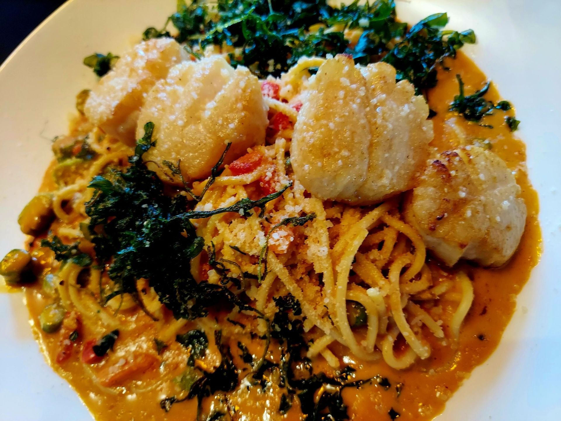Pan-seared scallops atop a bed of spaghetti in a creamy orange sauce, garnished with dark leafy greens.