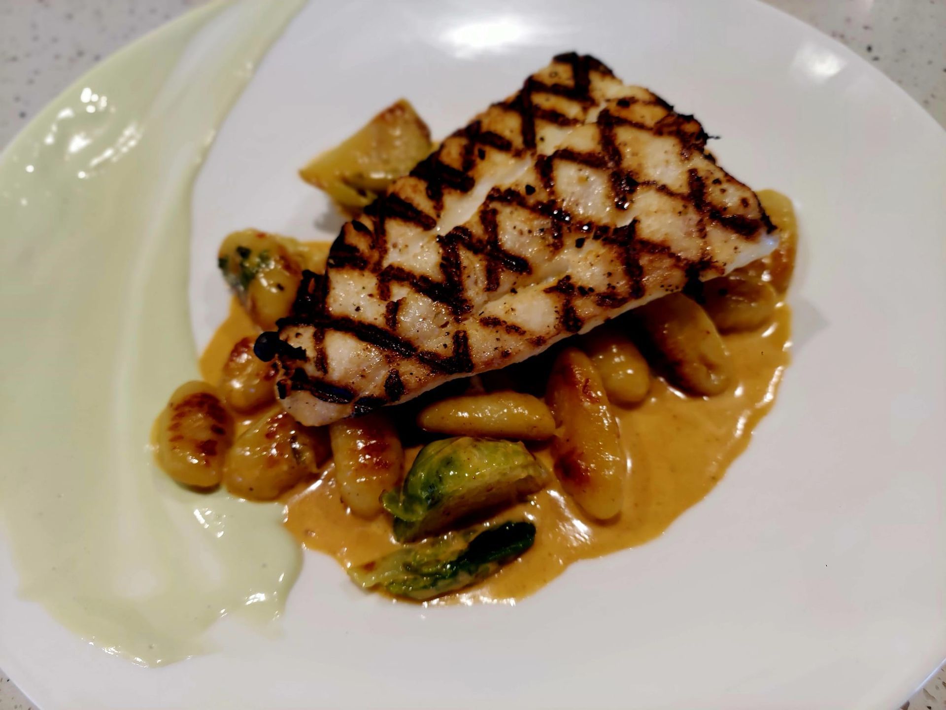 Grilled fish fillet served over gnocchi and roasted Brussels sprouts in a creamy orange sauce with a pale green puree.