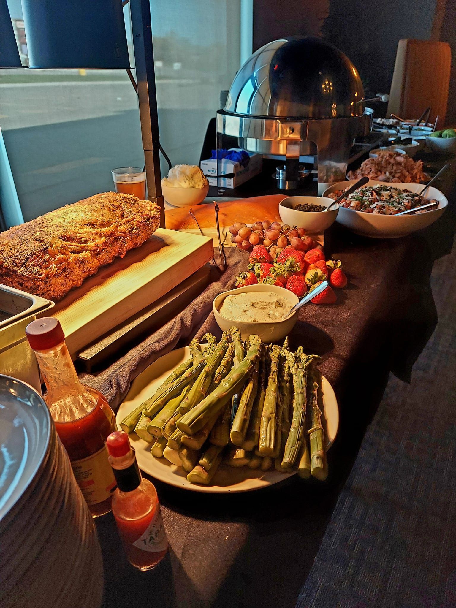 A buffet table features a loaf of bread, a platter of asparagus, bowls of salad, strawberries, and bottles of hot sauce.