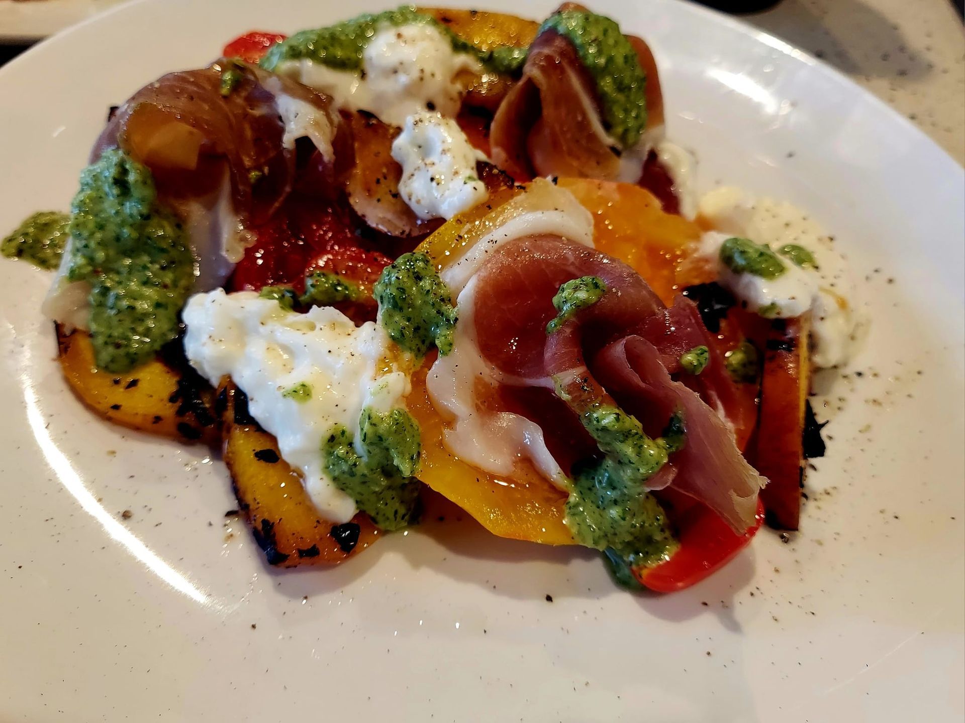 A white plate features grilled yellow peaches topped with ribbons of prosciutto, dollops of white cheese, and green pesto.