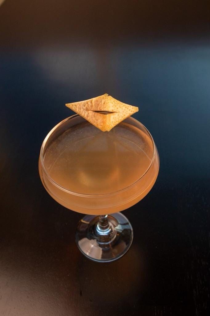A light amber cocktail in a stemmed coupe glass, garnished with a diamond-shaped citrus peel on a dark surface.