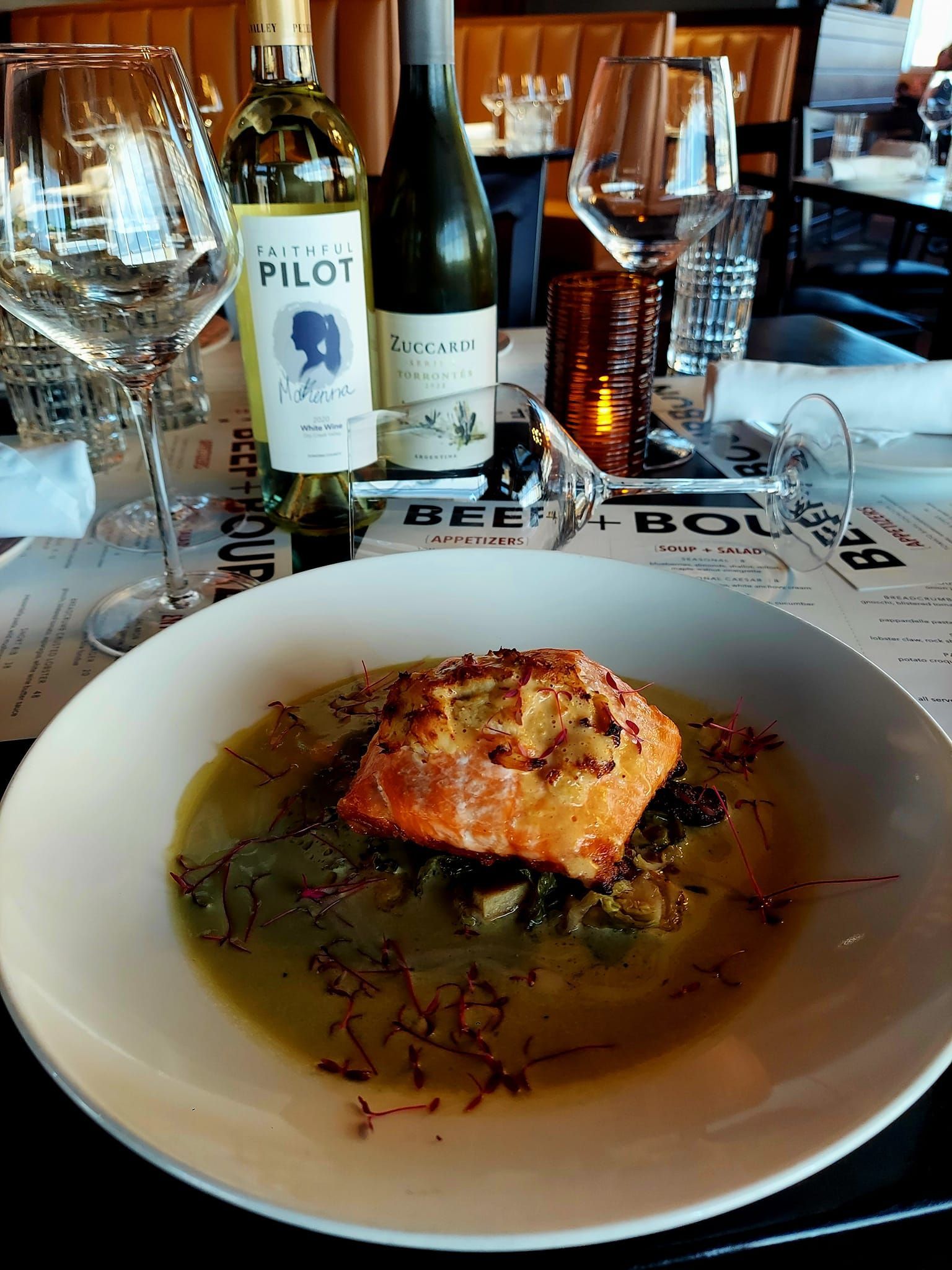 A crispy-skinned fillet of fish served in a shallow bowl of broth with garnish, on a table with wine bottles and glasses.
