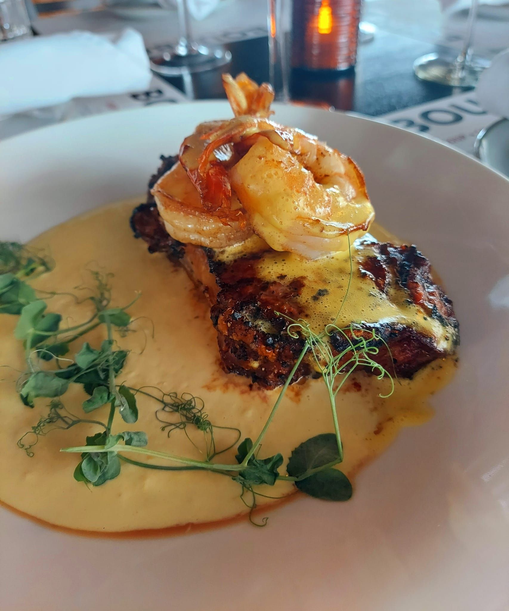 A seared steak topped with shrimp and yellow sauce, garnished with pea shoots on a white plate in a restaurant.