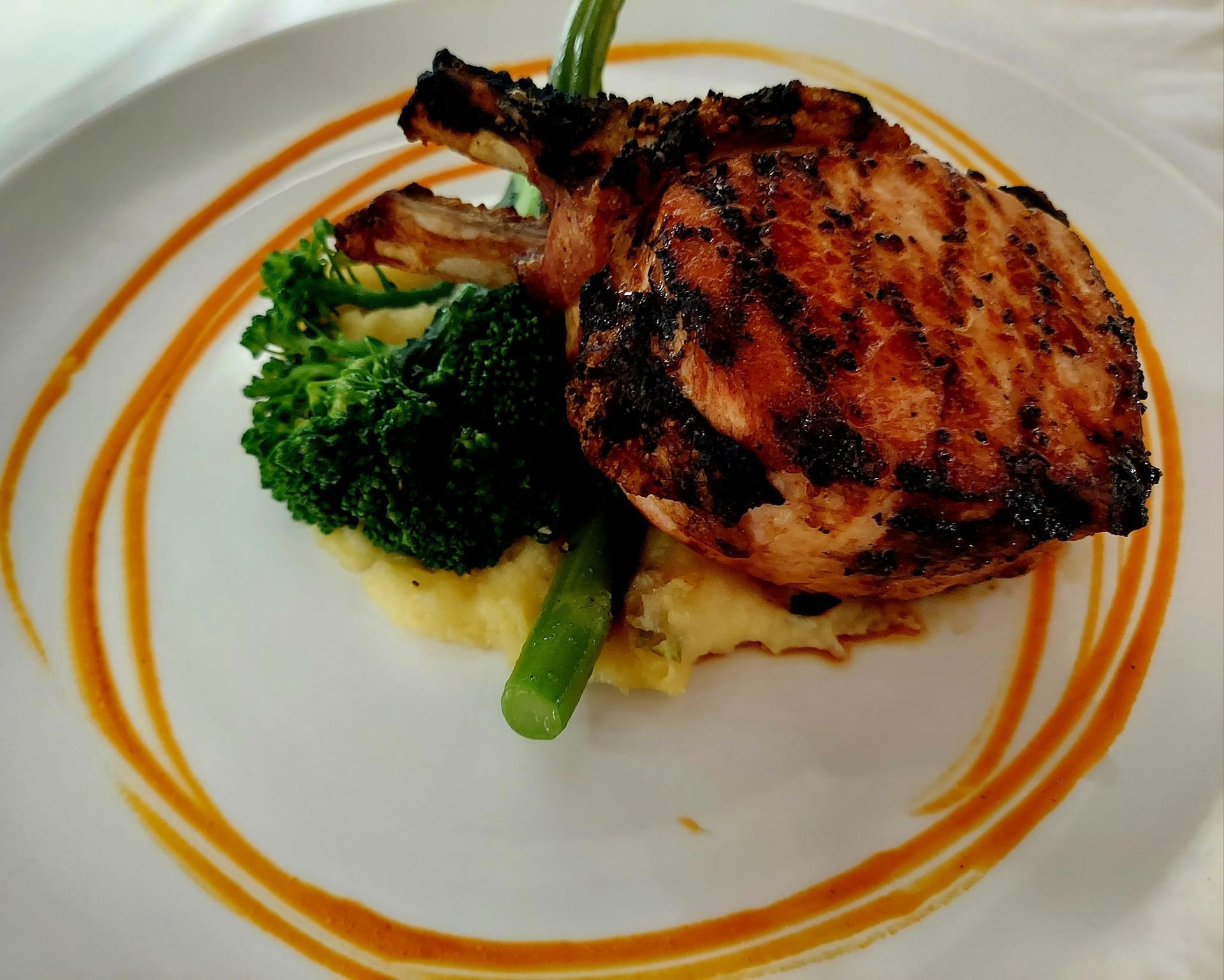 A grilled pork chop served with mashed potatoes, broccoli, and asparagus on a plate with an orange sauce swirl.