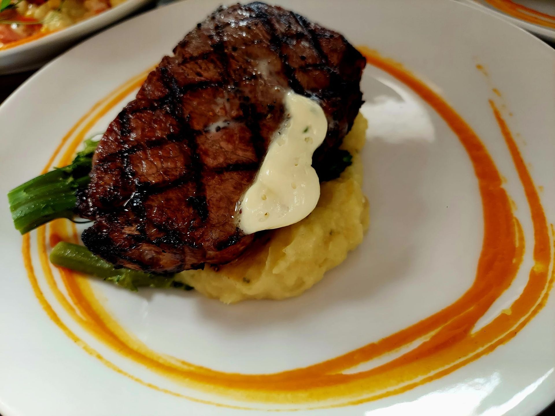 A grilled steak topped with a dollop of butter on a bed of mashed potatoes and green beans, plated with a sauce swirl.
