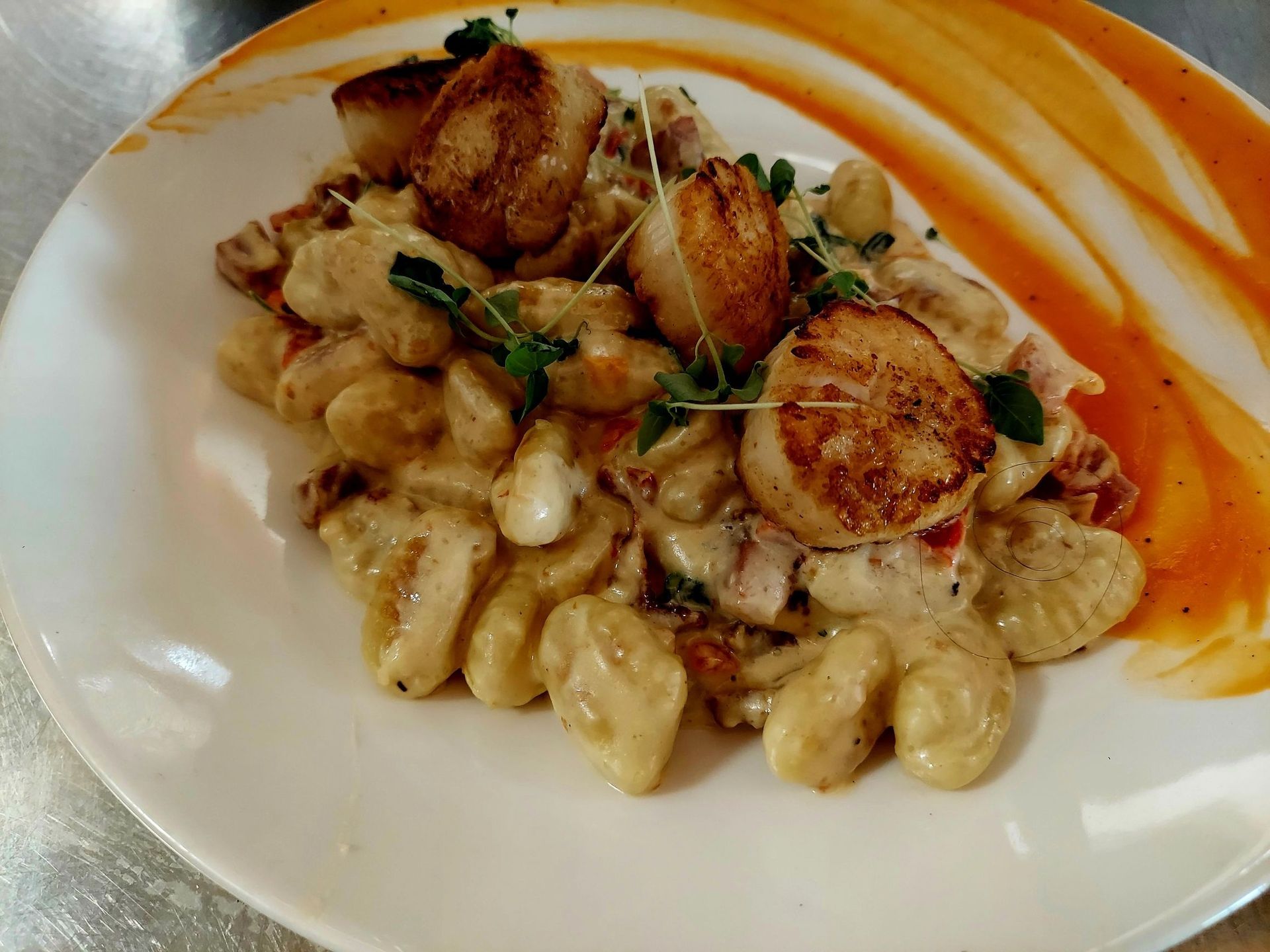 Three seared scallops atop a mound of creamy gnocchi, garnished with fresh herbs, served on a white plate with sauce.