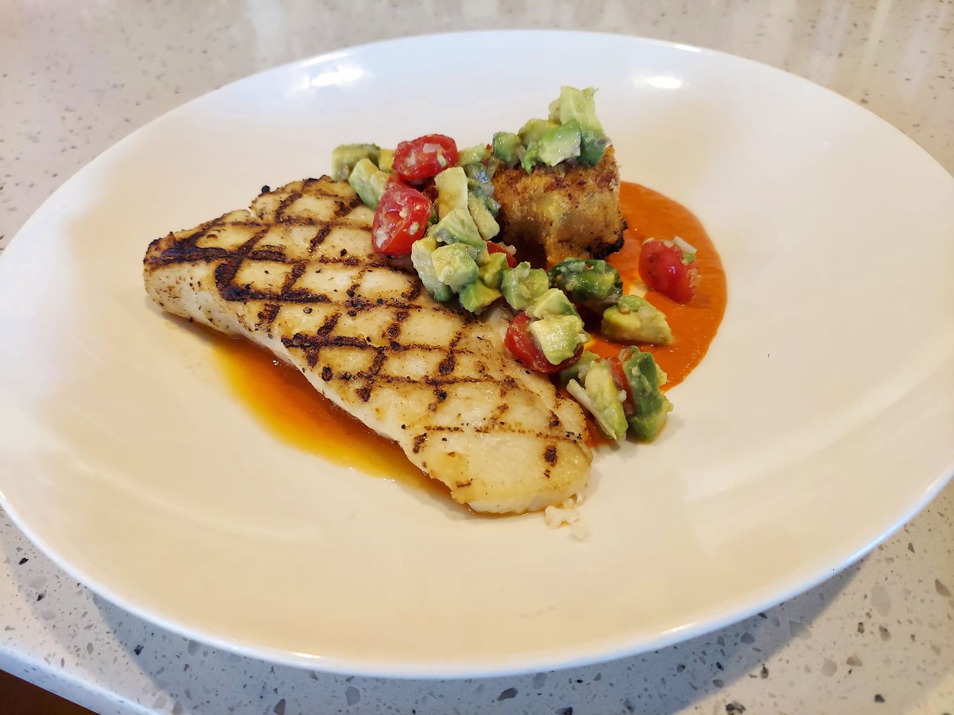 A grilled fish fillet topped with avocado and tomato salsa, served on a plate with a vibrant orange sauce.