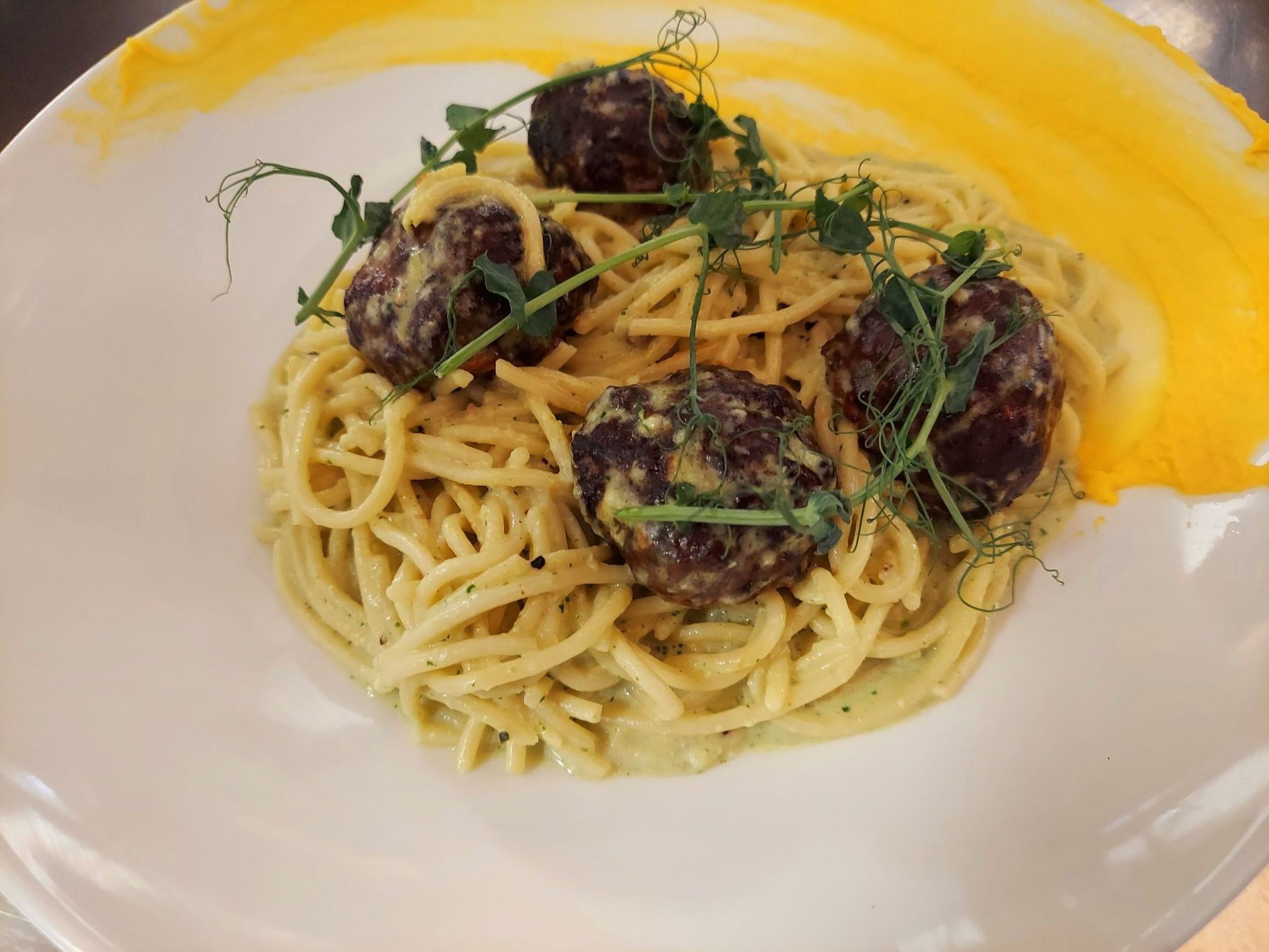 A plate of spaghetti with four meatballs, topped with microgreens and a decorative yellow sauce streak.