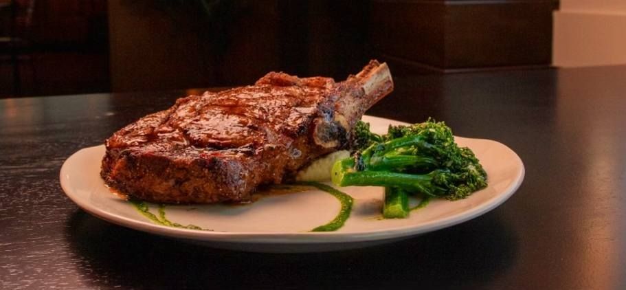 A bone-in ribeye steak served with a side of green broccolini on a white plate against a dark background.