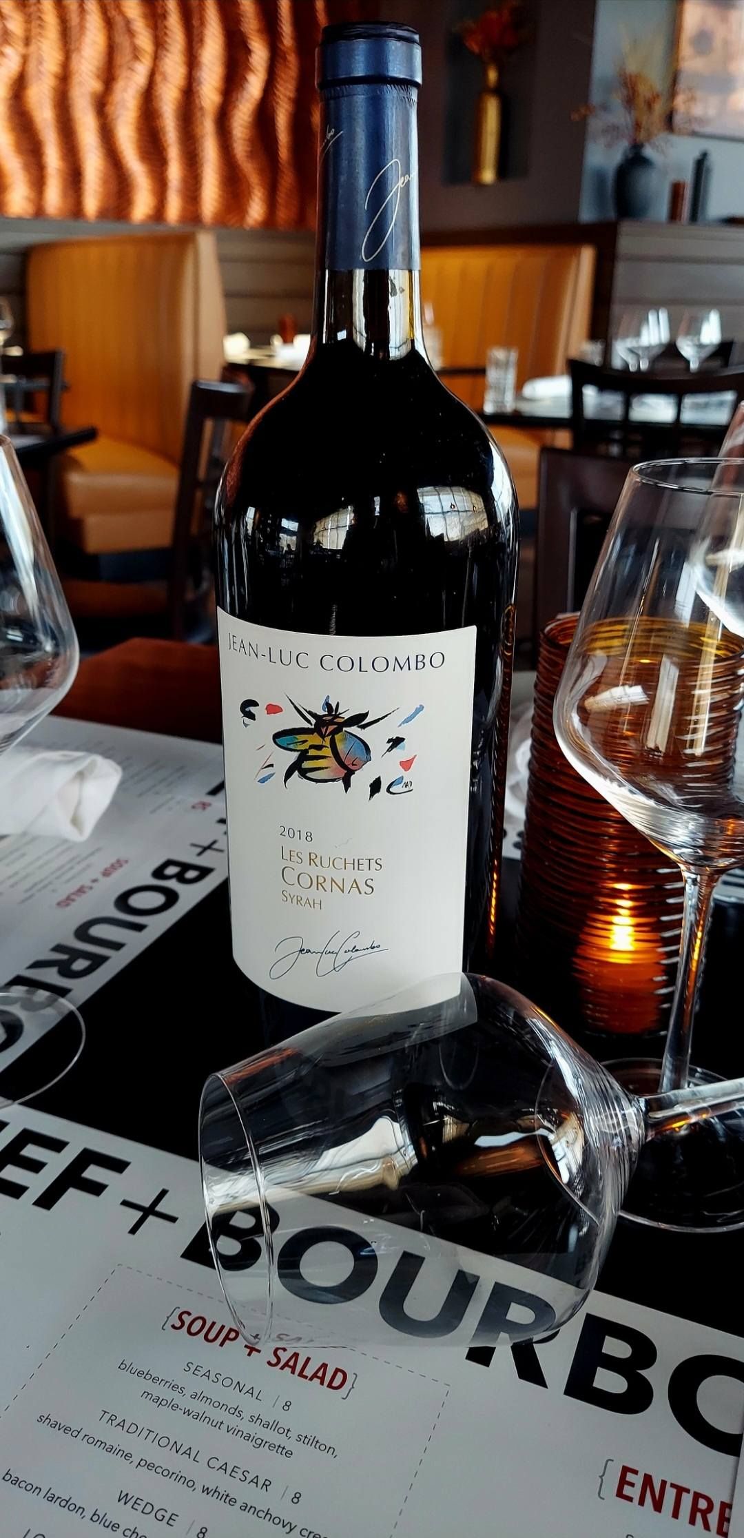A wine bottle with a colorful illustration on the label sits on a restaurant table next to wine glasses and a menu.
