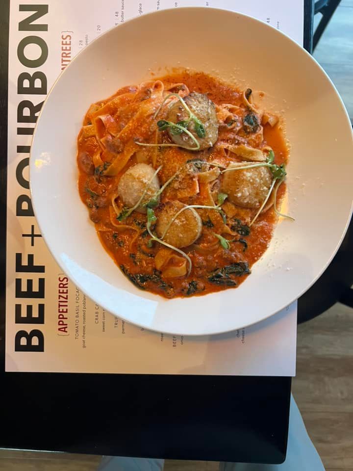 A plate of pasta with seared scallops in a red sauce, served on a table next to a Beef + Bourbon restaurant menu.
