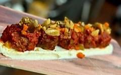 A row of cooked meatballs topped with tomato sauce and chopped vegetables, resting on a bed of white cream on a tray.