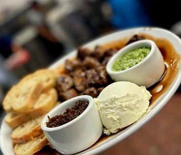 A platter with toasted bread, sautéed mushrooms, savory jam, green herb sauce, and a scoop of white spread or cheese.