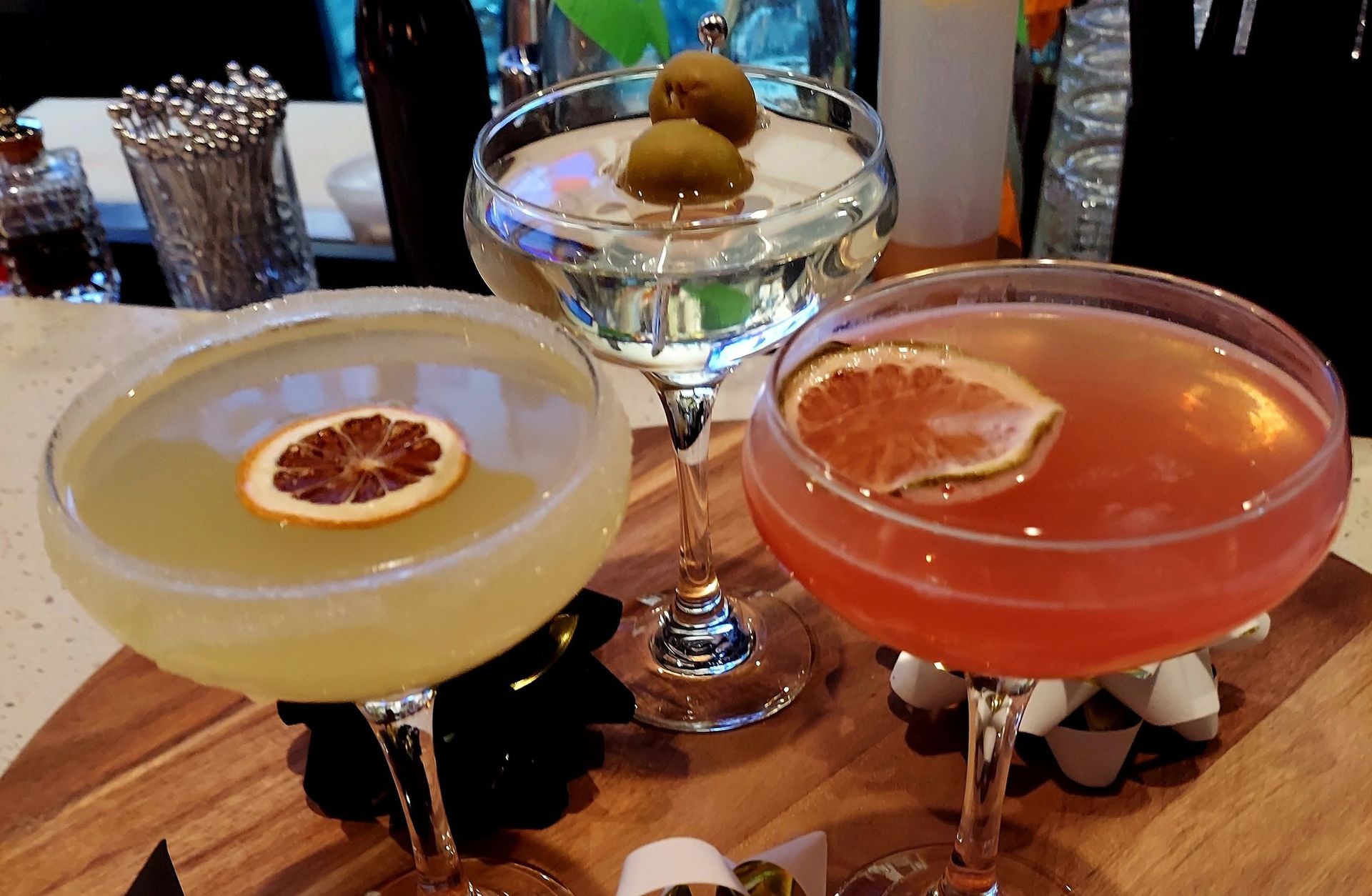 Three cocktails, one clear with two olives and two with citrus slices, served on a wooden board at a bar.