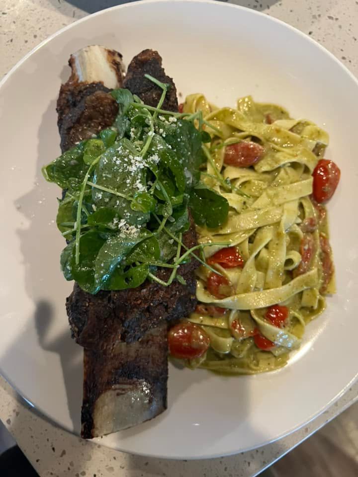A large braised beef rib topped with fresh arugula on a white plate next to a serving of fettuccine pasta with tomatoes.