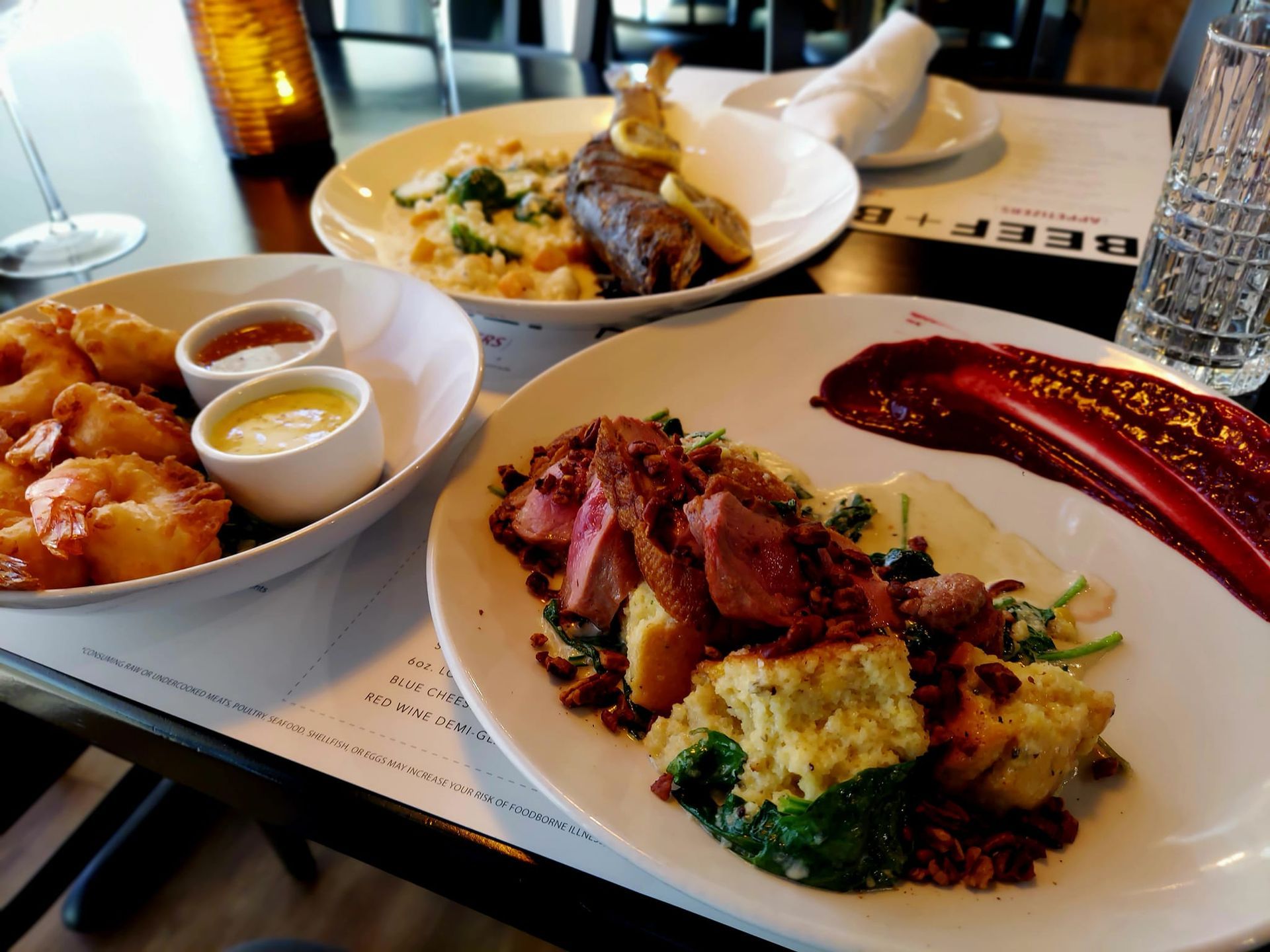 Three plates of gourmet dishes, including shrimp and meat, served on a table with a menu in a restaurant setting.