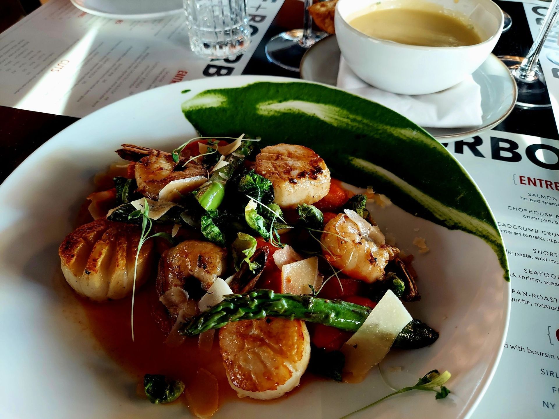 A close-up of a plated scallop and shrimp dish with asparagus and cheese, served with a side of soup at a restaurant.