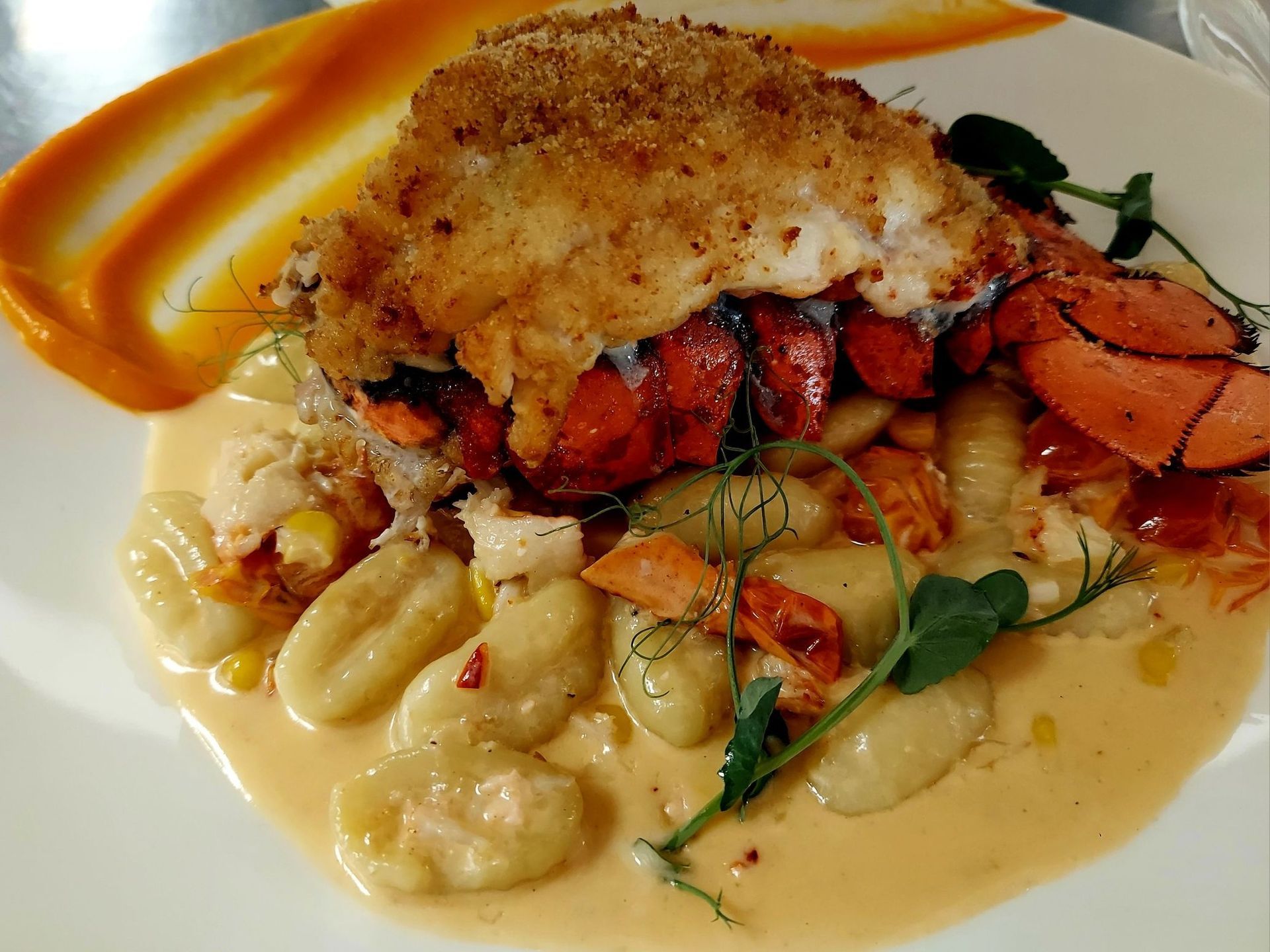 A breaded lobster tail served over gnocchi in a creamy sauce, garnished with herbs and a swirl of orange puree on a plate.