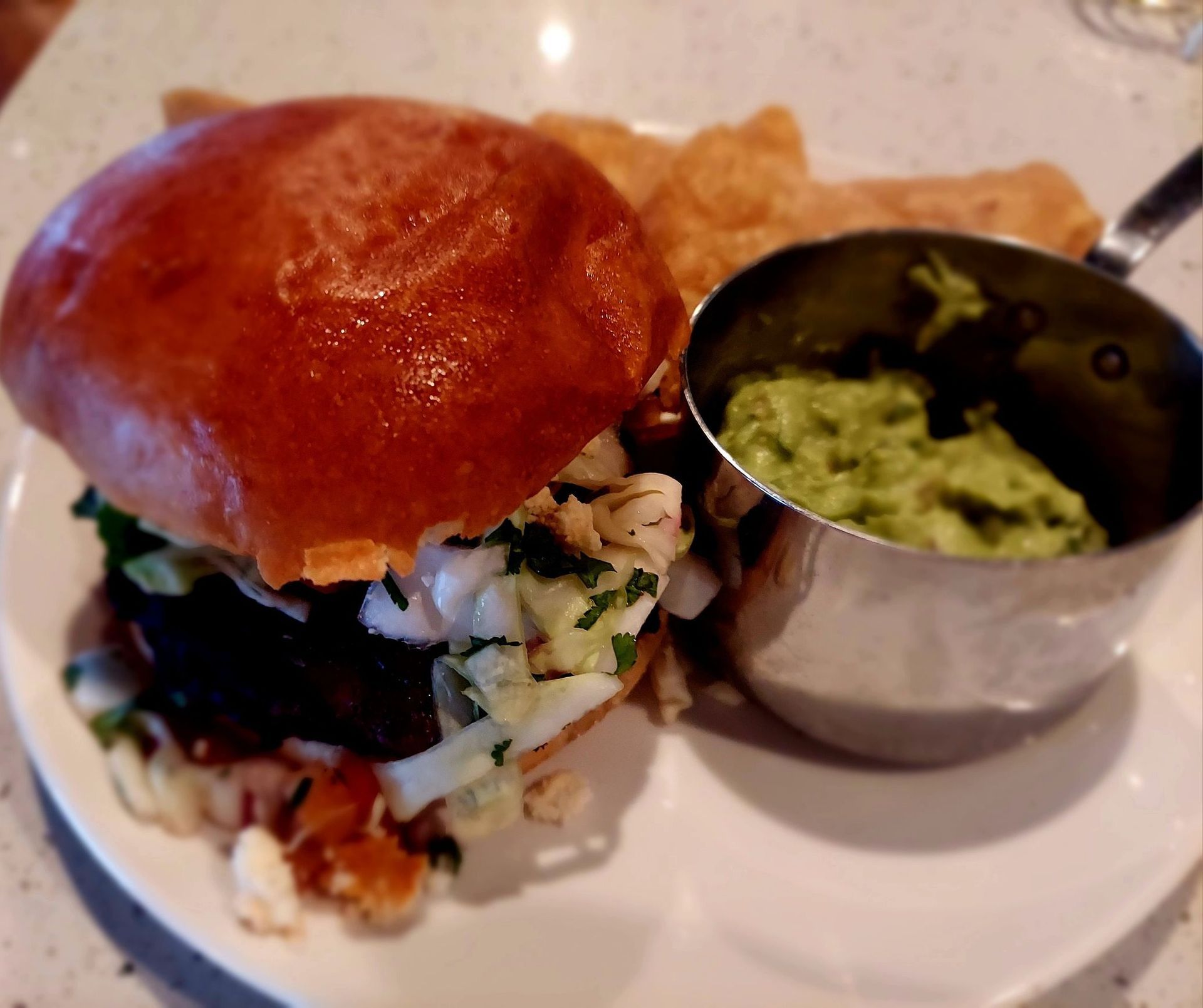 A brioche bun sandwich topped with slaw next to a small metal cup of green dip and side of chips on a white plate.