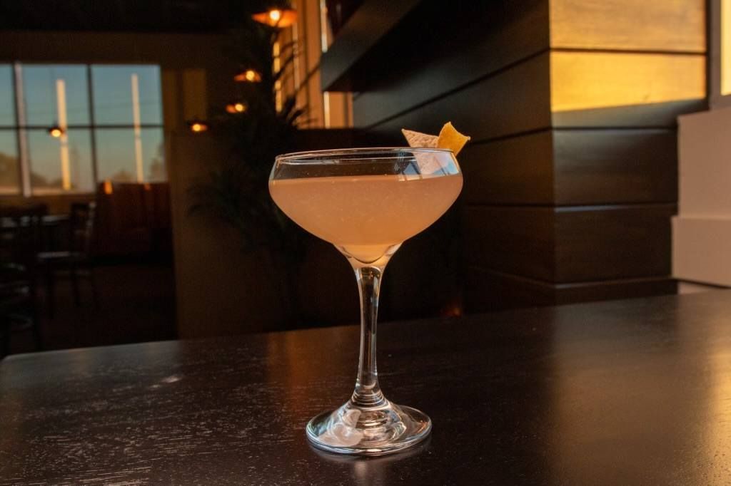 A stemmed cocktail glass filled with a pale yellow drink, garnished with a citrus peel, sitting on a dark table at sunset.