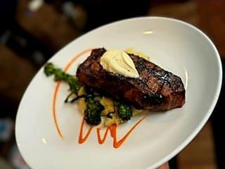 A grilled steak topped with a pat of butter, served on a bed of greens with decorative red sauce swirls on a white plate.