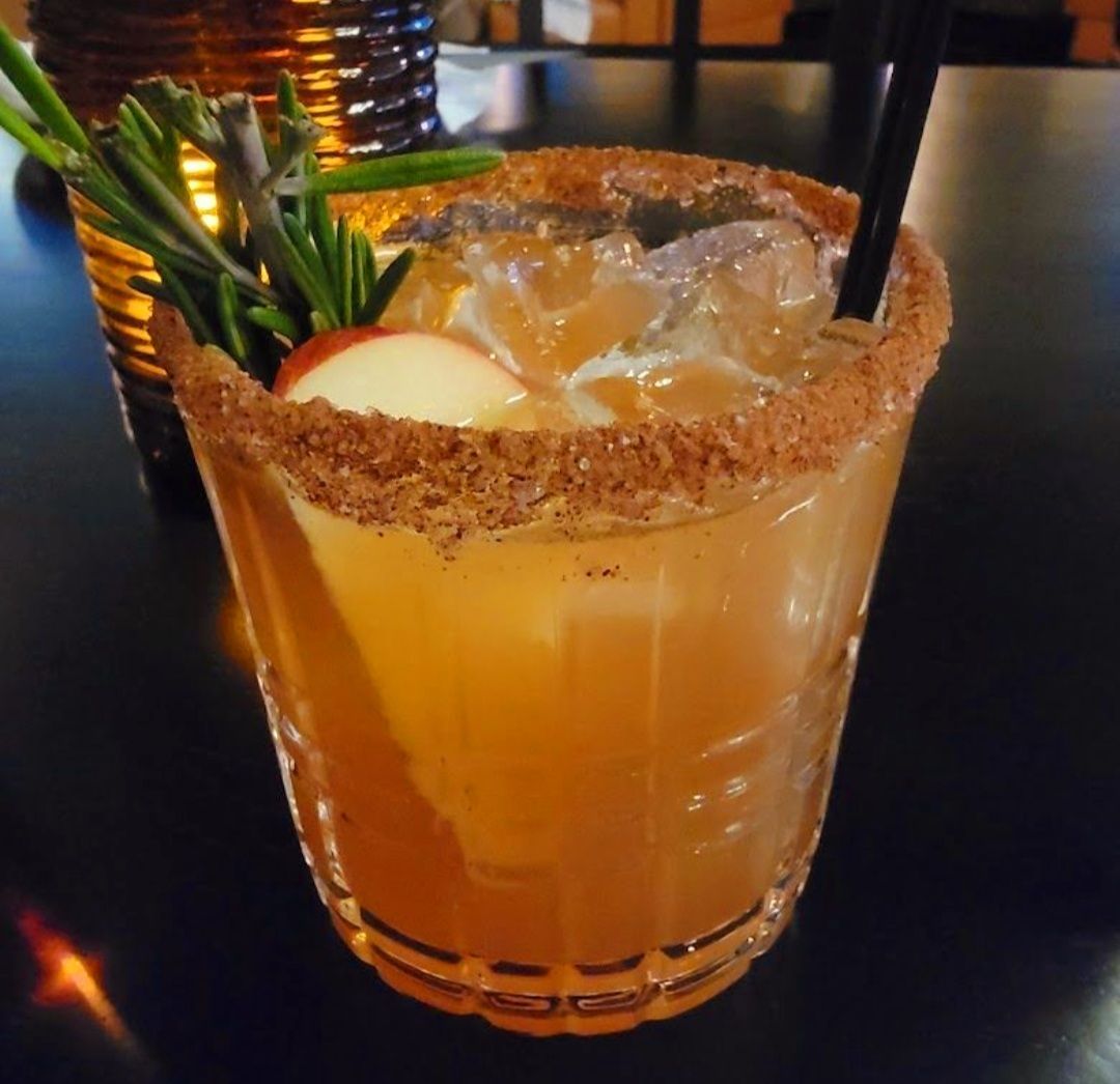 A glass cocktail with a cinnamon-sugar rim, ice, apple slice garnish, and a sprig of rosemary on a dark surface.