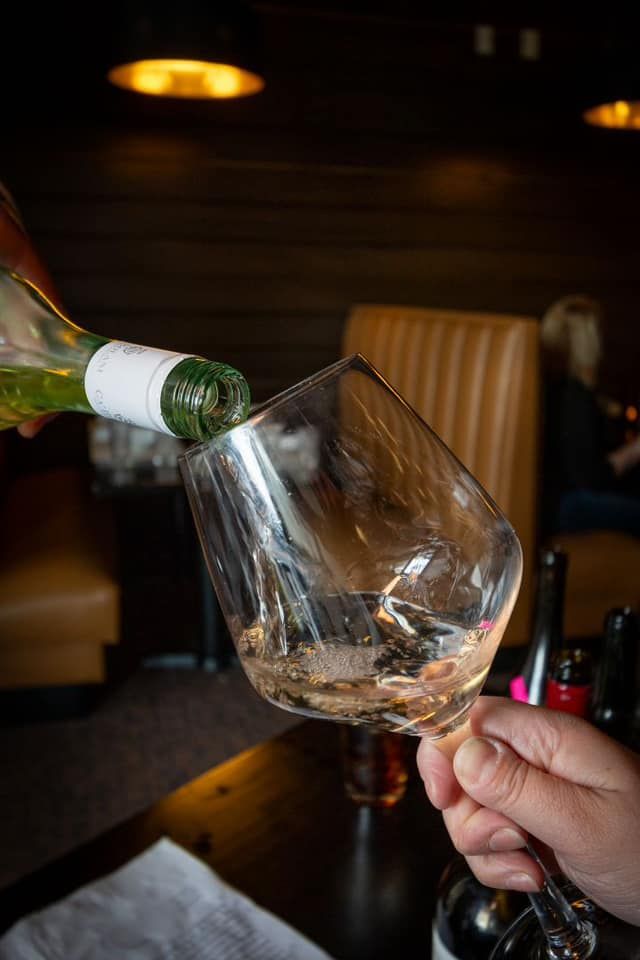 A person holds a wine glass as white wine is being poured into it from a glass bottle in a dimly lit restaurant booth.