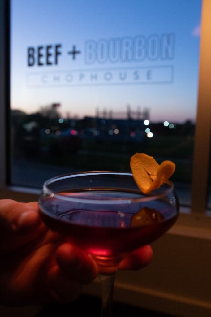 A hand holds a coupe glass filled with a red cocktail, garnished with a citrus twist, at Beef + Bourbon Chophouse.