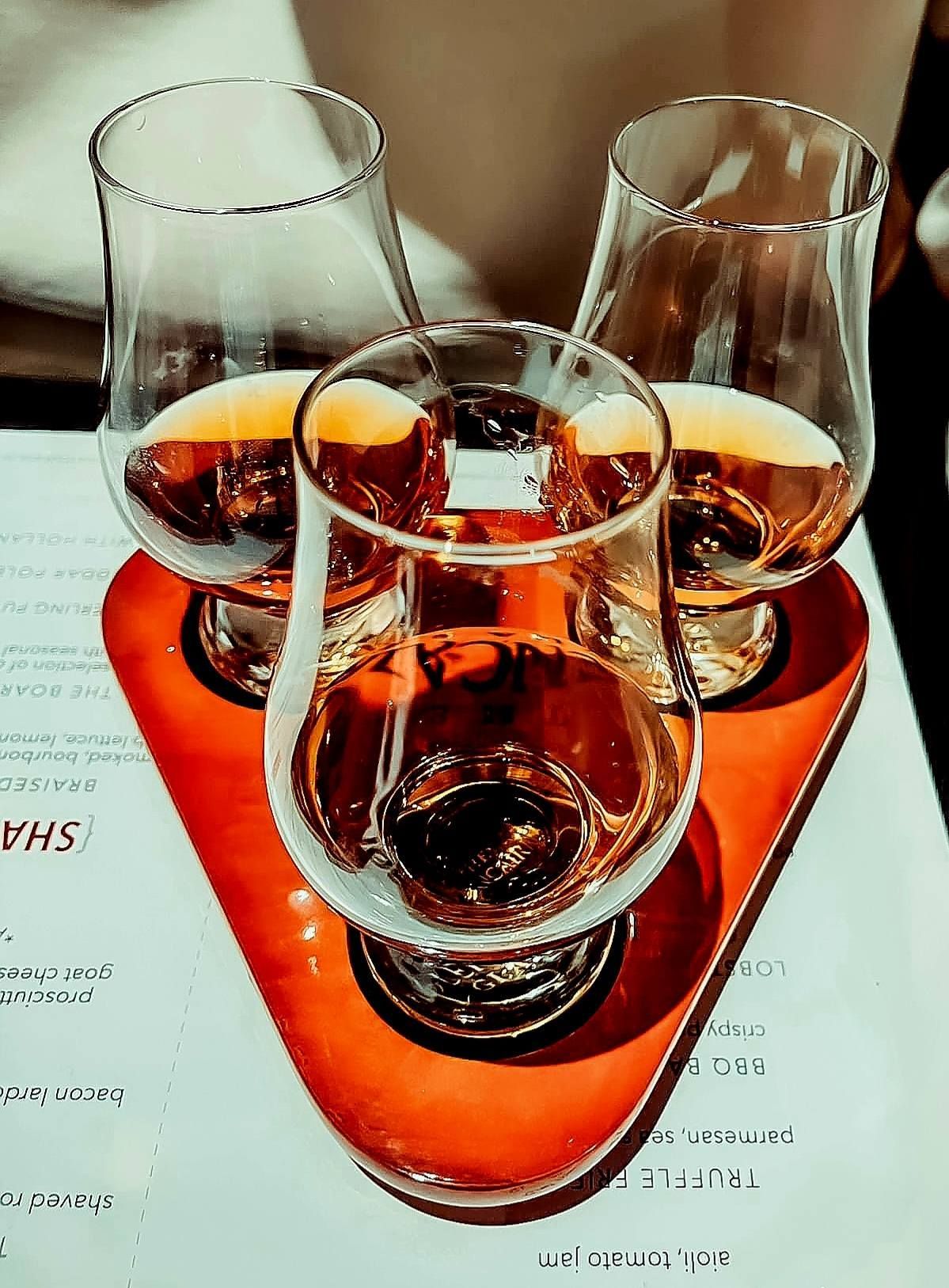 A tasting flight of three amber-colored whiskeys in tulip-shaped glasses resting on a triangular orange paddle board.