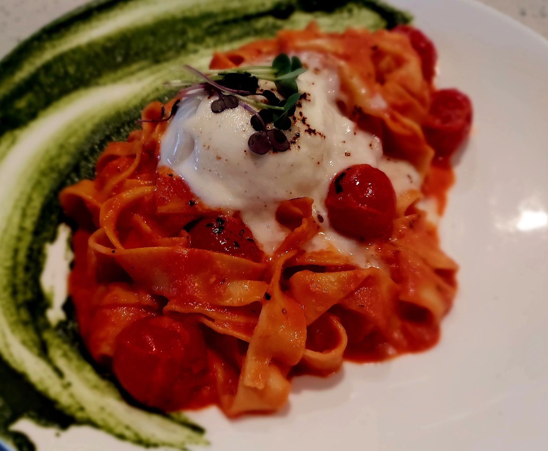 A mound of red tomato pasta topped with burrata cheese and microgreens, served alongside a streak of green herb sauce.