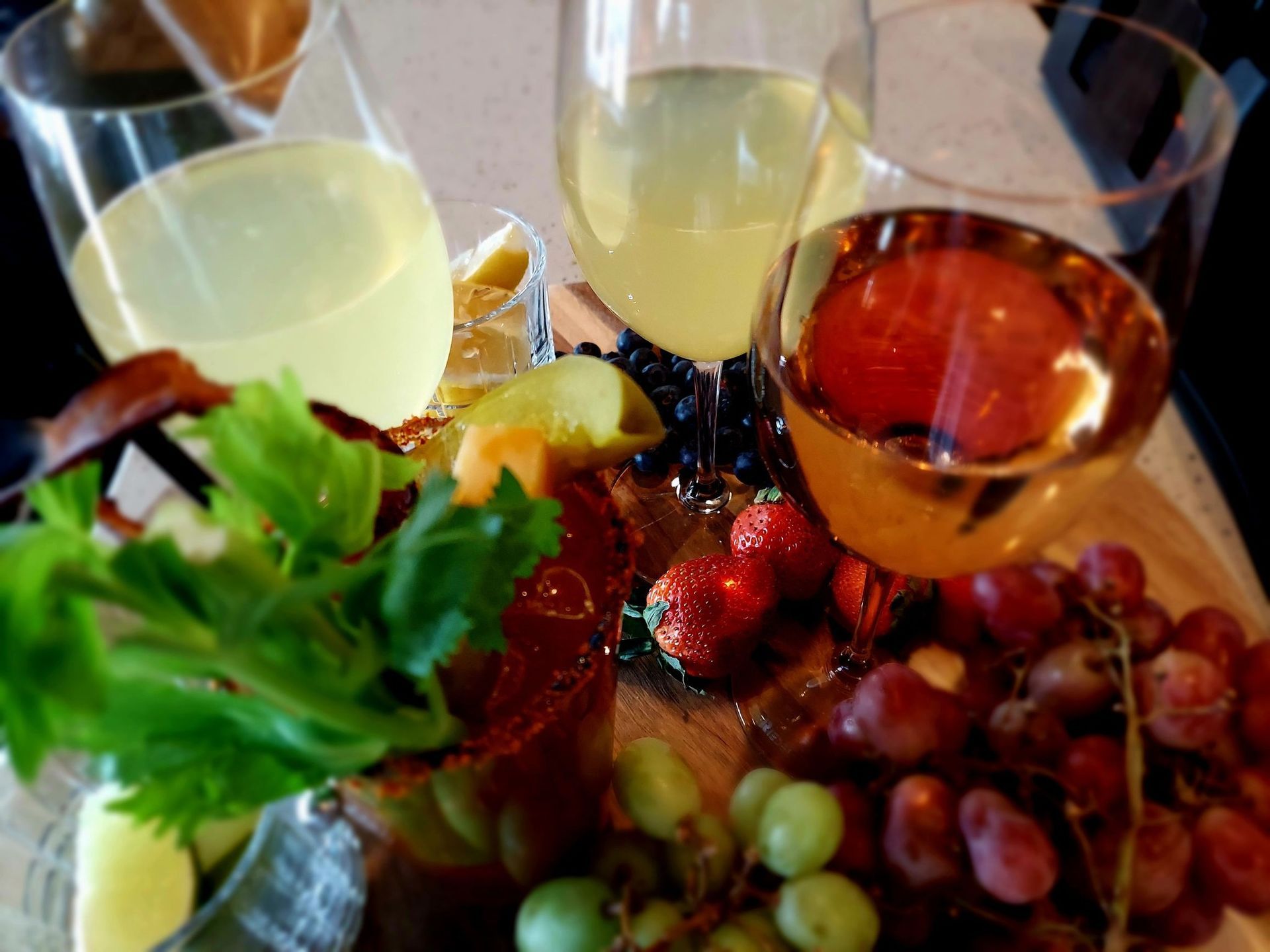 A charcuterie board with a Bloody Mary, wine, grapes, strawberries, and celery on a light surface.