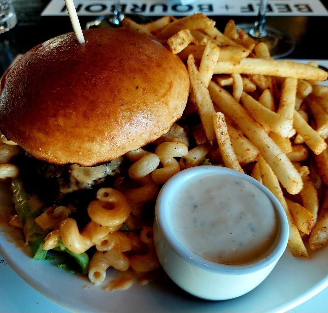 A hamburger topped with mac and cheese, served with a side of fries and dipping sauce on a white plate.