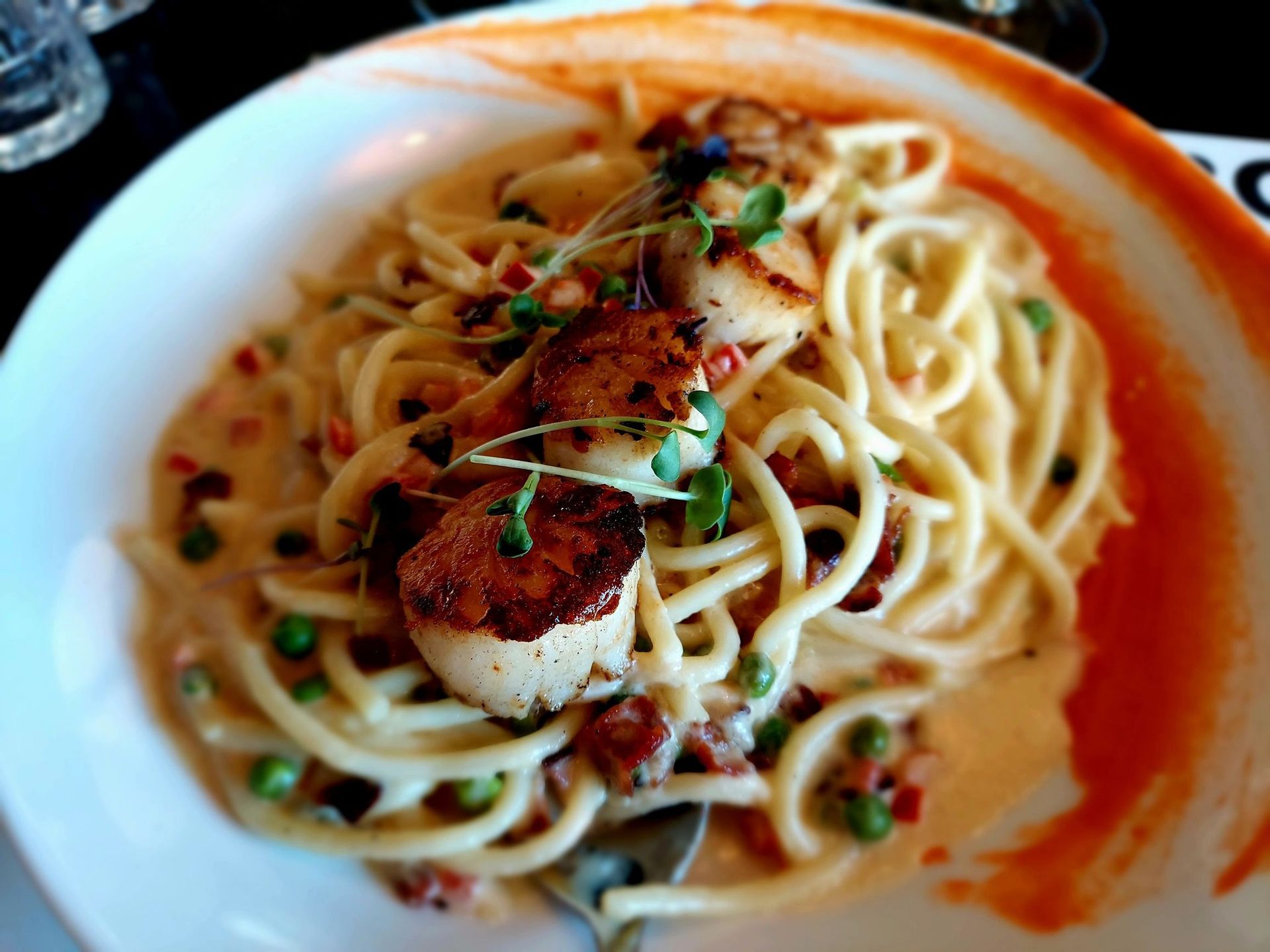 A white bowl of pasta topped with three pan-seared scallops and peas in a creamy, orange-red sauce.