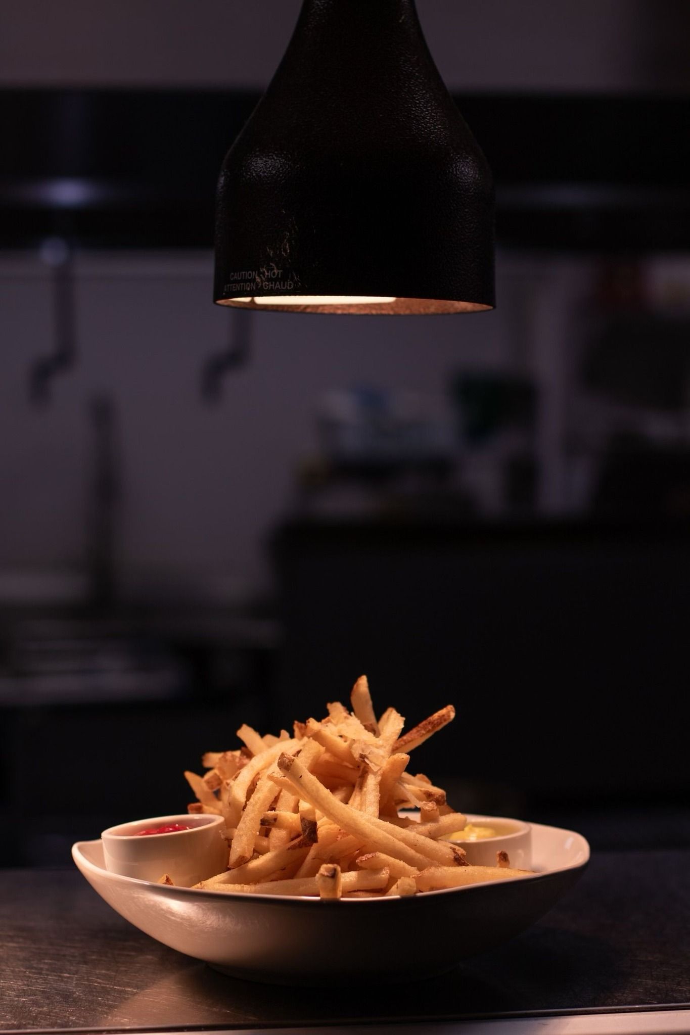 A bowl of golden french fries sits on a dark surface under a single hanging light in a dimly lit setting.