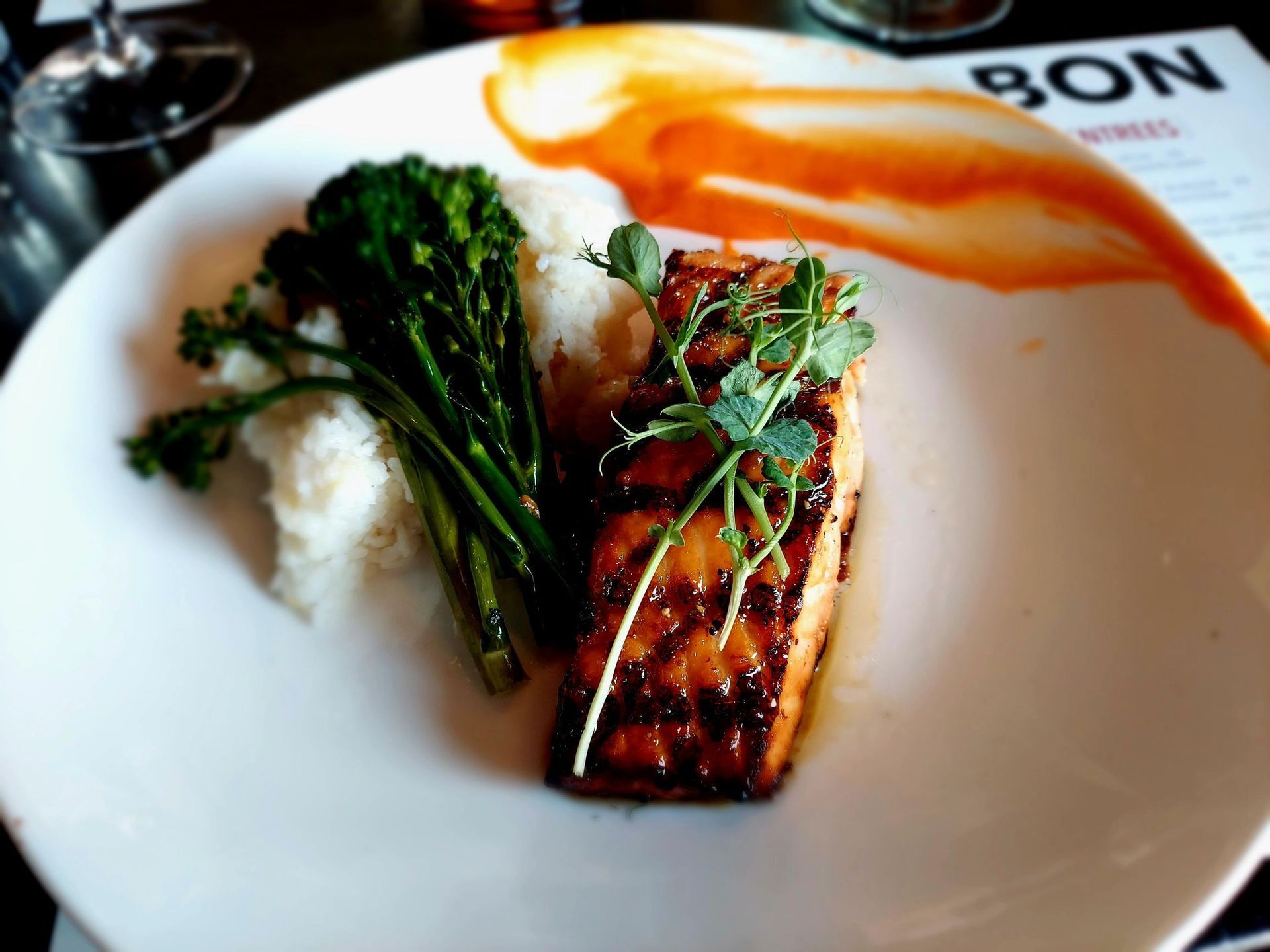 A glazed salmon fillet with rice and broccoli, garnished with sprouts on a white plate with a decorative sauce streak.