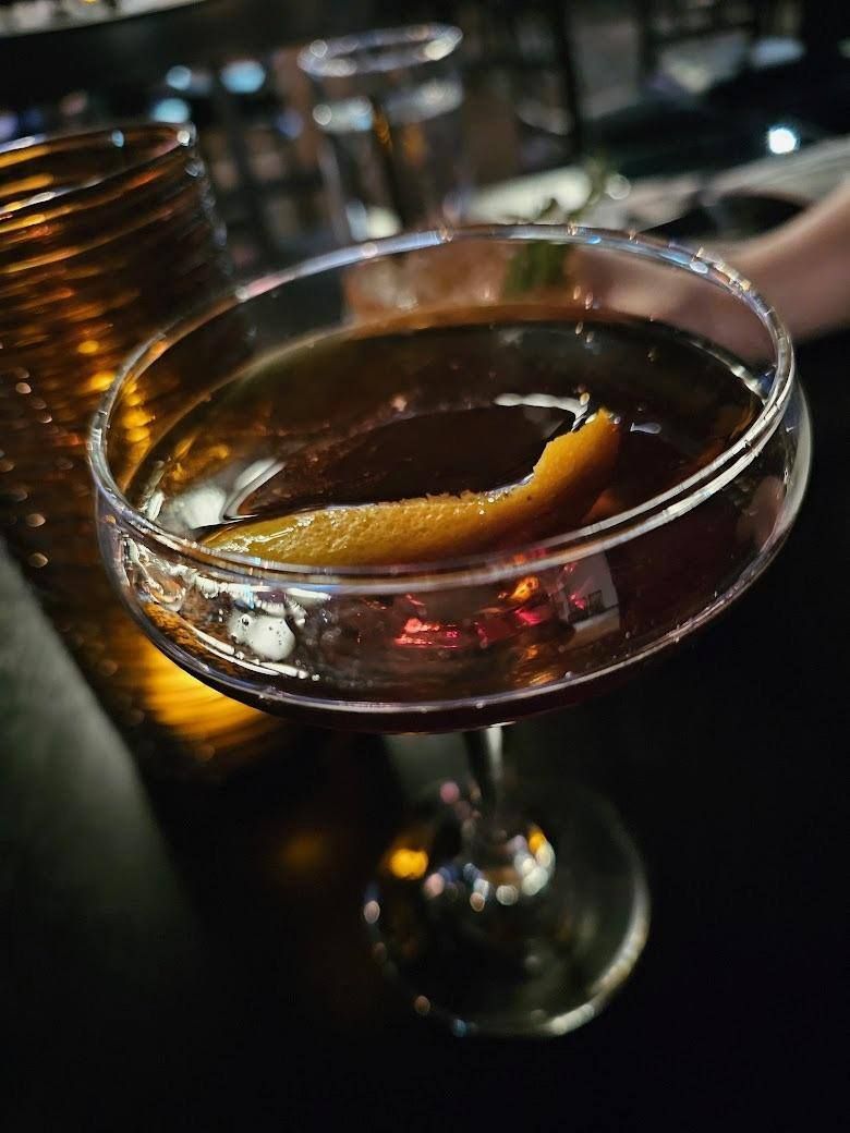 A coupe glass filled with a dark amber cocktail, garnished with an orange twist, sitting on a dark surface.