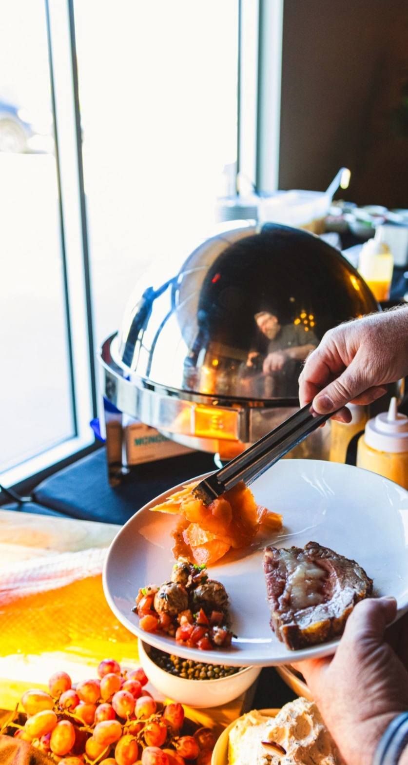 A person uses tongs to add food to a plate at a buffet, with a metal warmer and bowls of fresh fruit nearby.