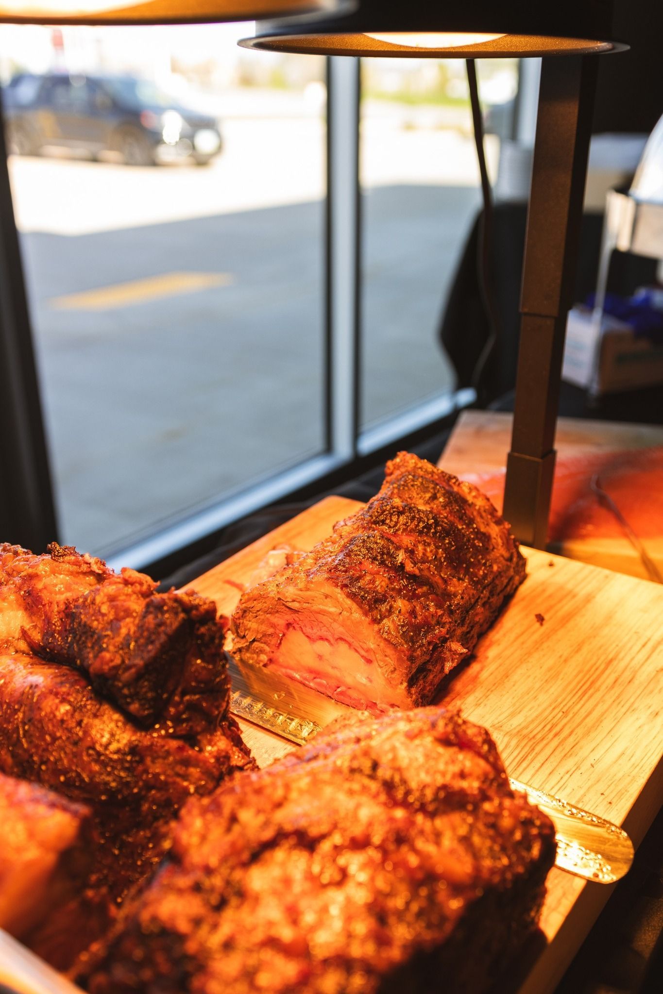 Several cuts of roasted, seasoned meat sit on a wooden serving board under a warm lamp near a window.