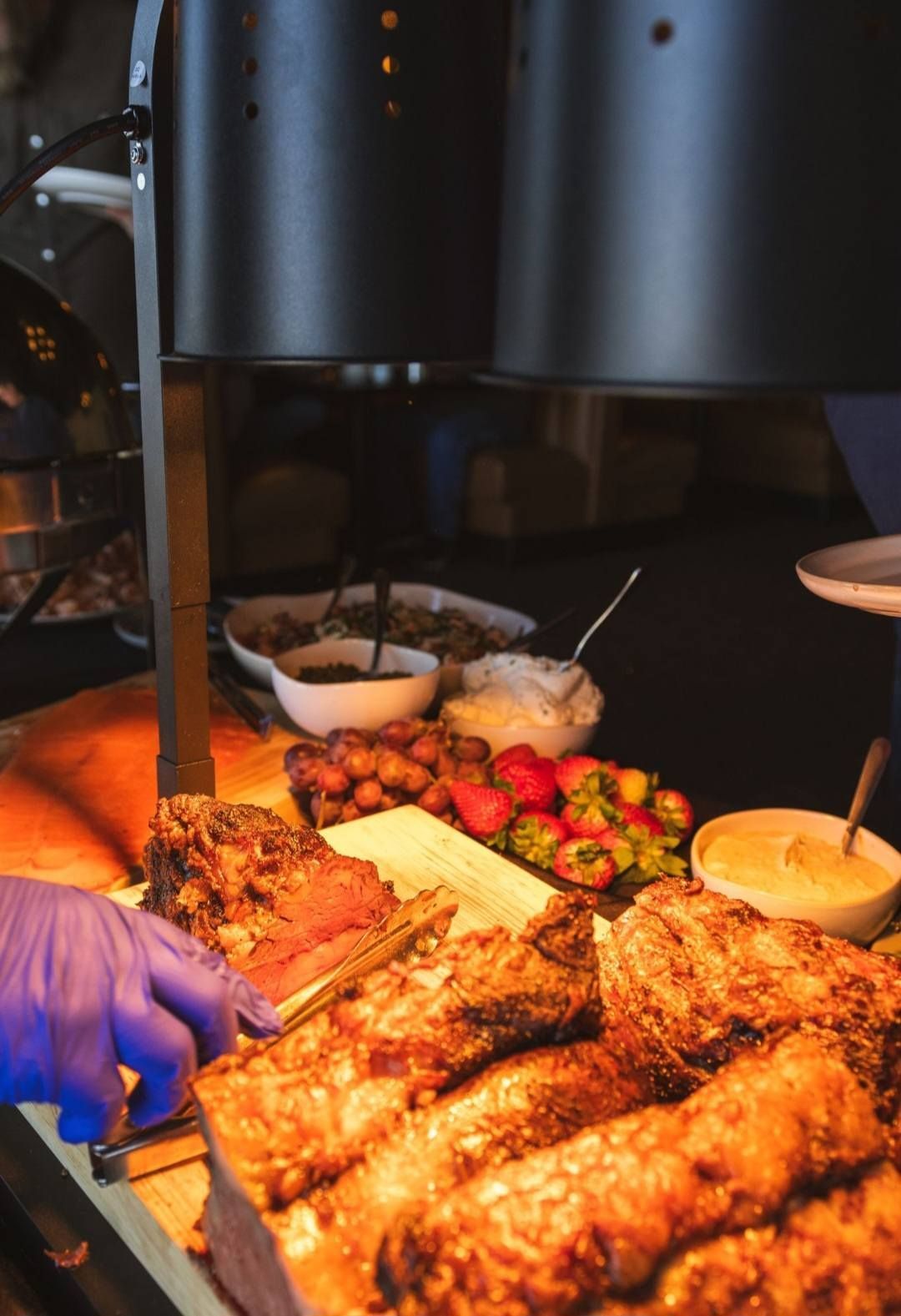 A gloved hand carves roast meat at a buffet line featuring sides, berries, and sauces under warm heat lamps.