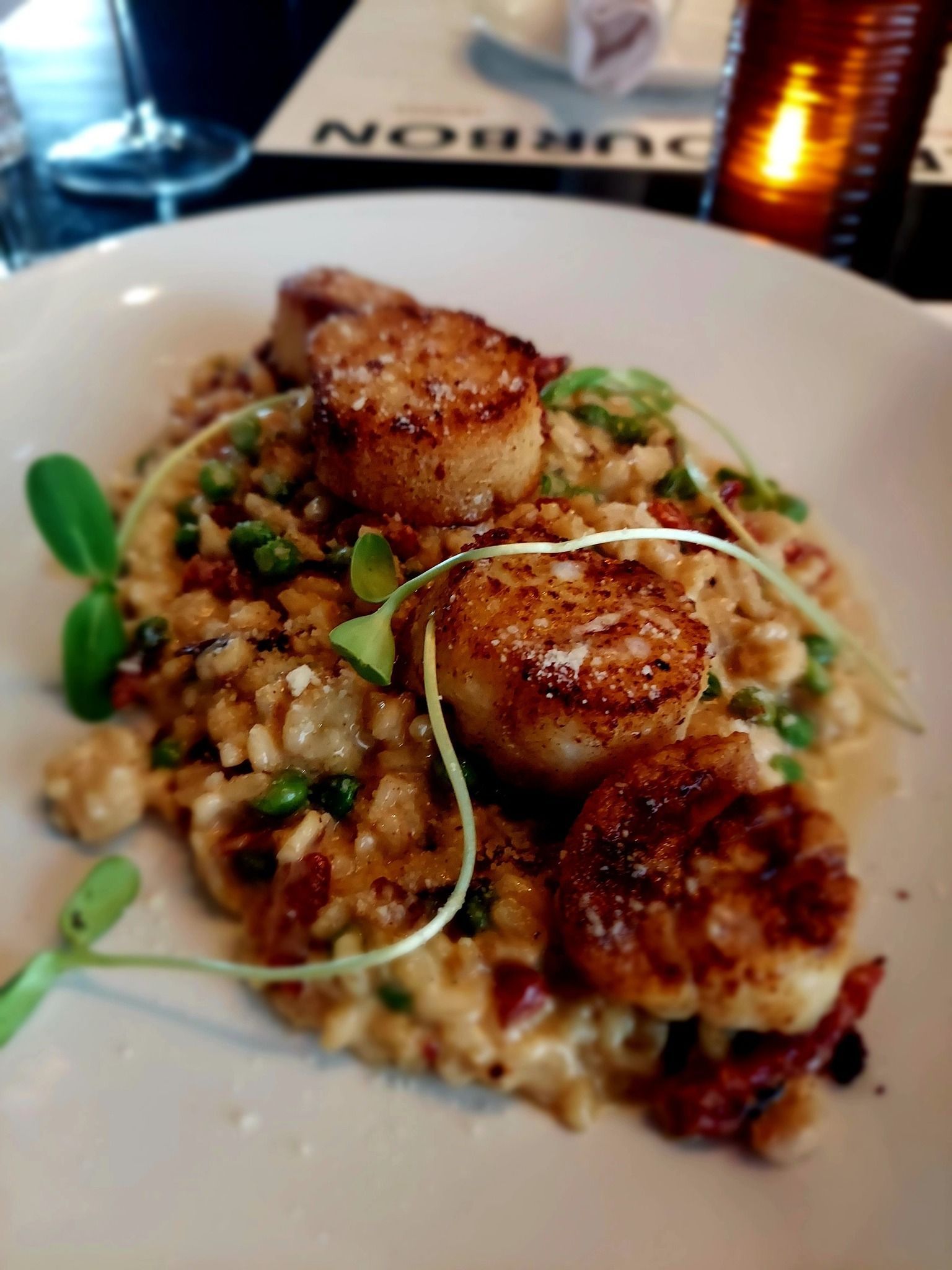 A plate of seared scallops served over a creamy grain dish with green peas, garnished with microgreens.