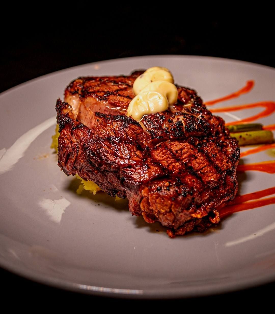 A grilled steak topped with a dollop of butter sits on a white plate with green asparagus and streaks of red sauce.