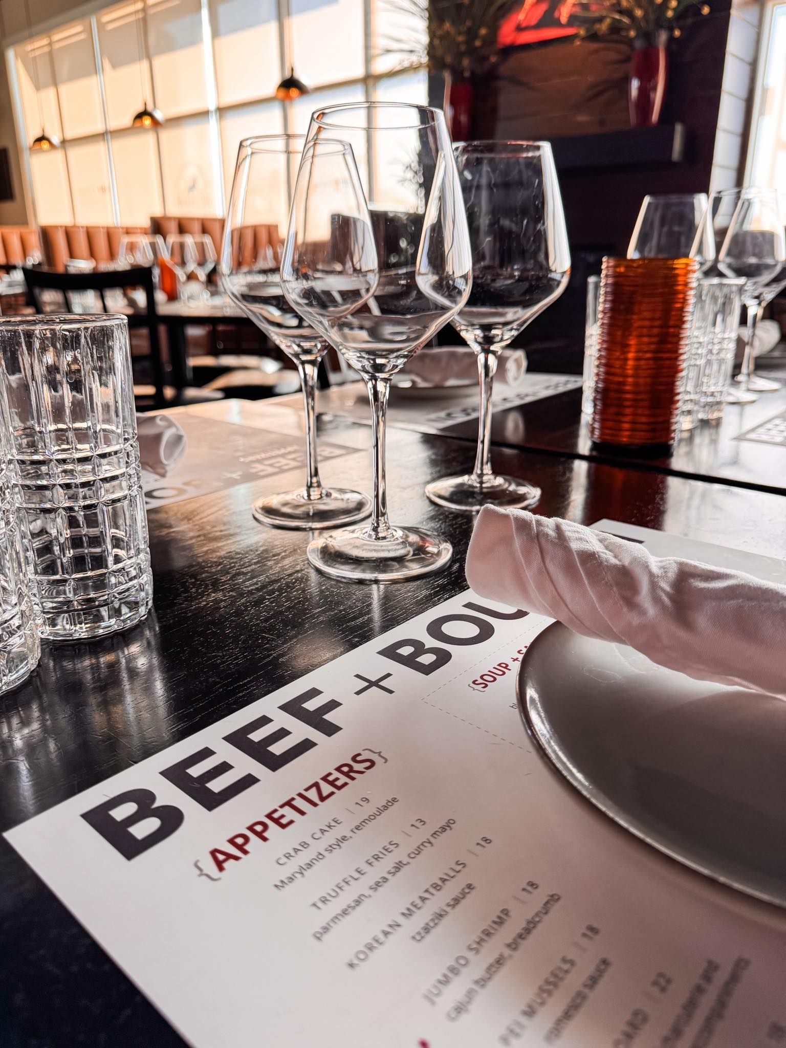A restaurant table set with wine glasses, a white napkin, and a Beef + Bottle menu featuring appetizers.