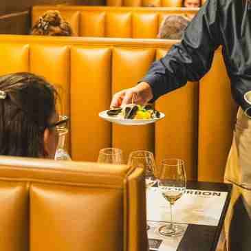 A server holds a small plate of food for a customer seated in a tan, tufted-back booth in a restaurant.