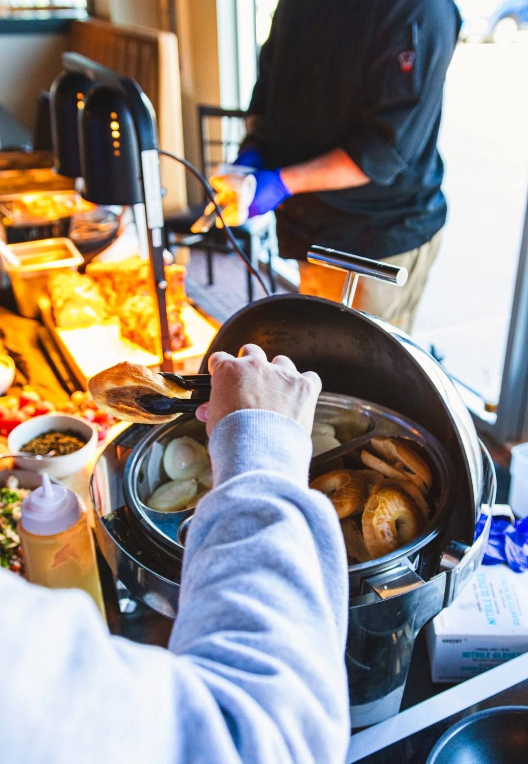 A person in a light hoodie uses tongs to serve bagels from a warming station in a buffet setting.