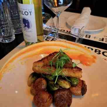 A plated gourmet meal featuring seared scallops and brussels sprouts, with a wine bottle in the background on a table.